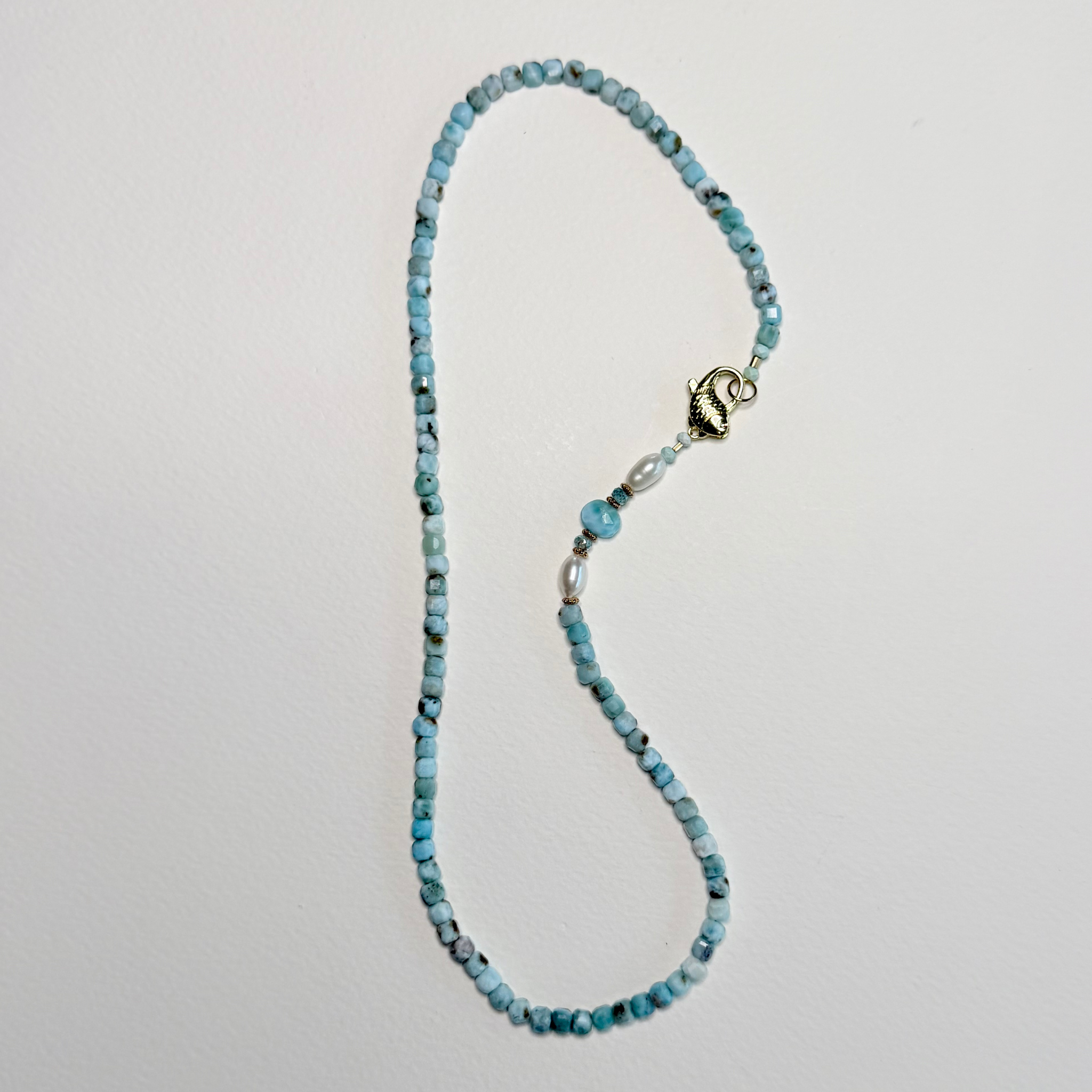 A beaded necklace made of small turquoise-blue and white marbled beads, featuring a silver-toned lobster clasp and a decorative silver spacer bead, laid flat on a light gray background.