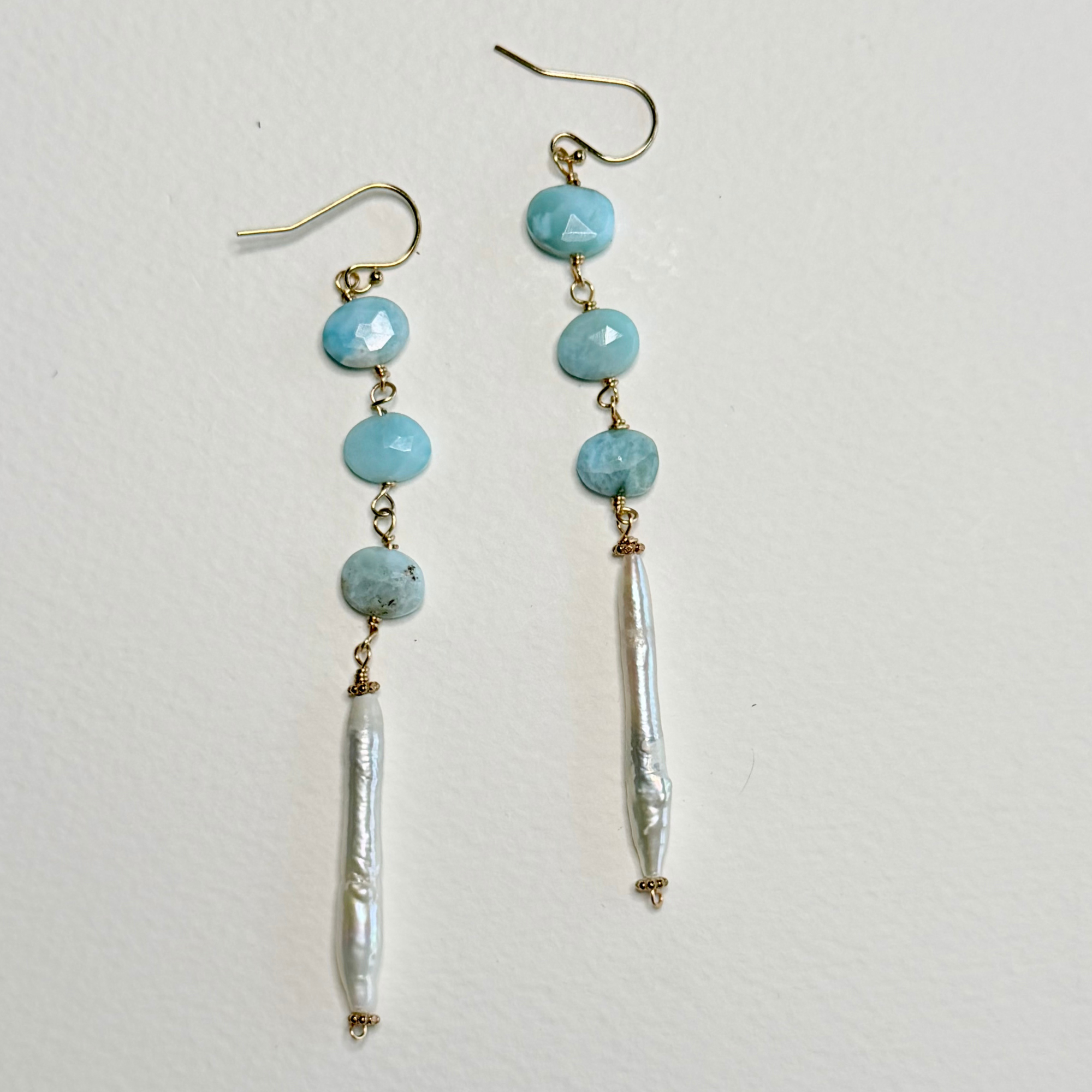 A pair of handmade dangle earrings featuring light blue larimar faceted beads, gold-tone hooks and connectors, and a long white baroque pearl drop, displayed on a textured off-white background.
