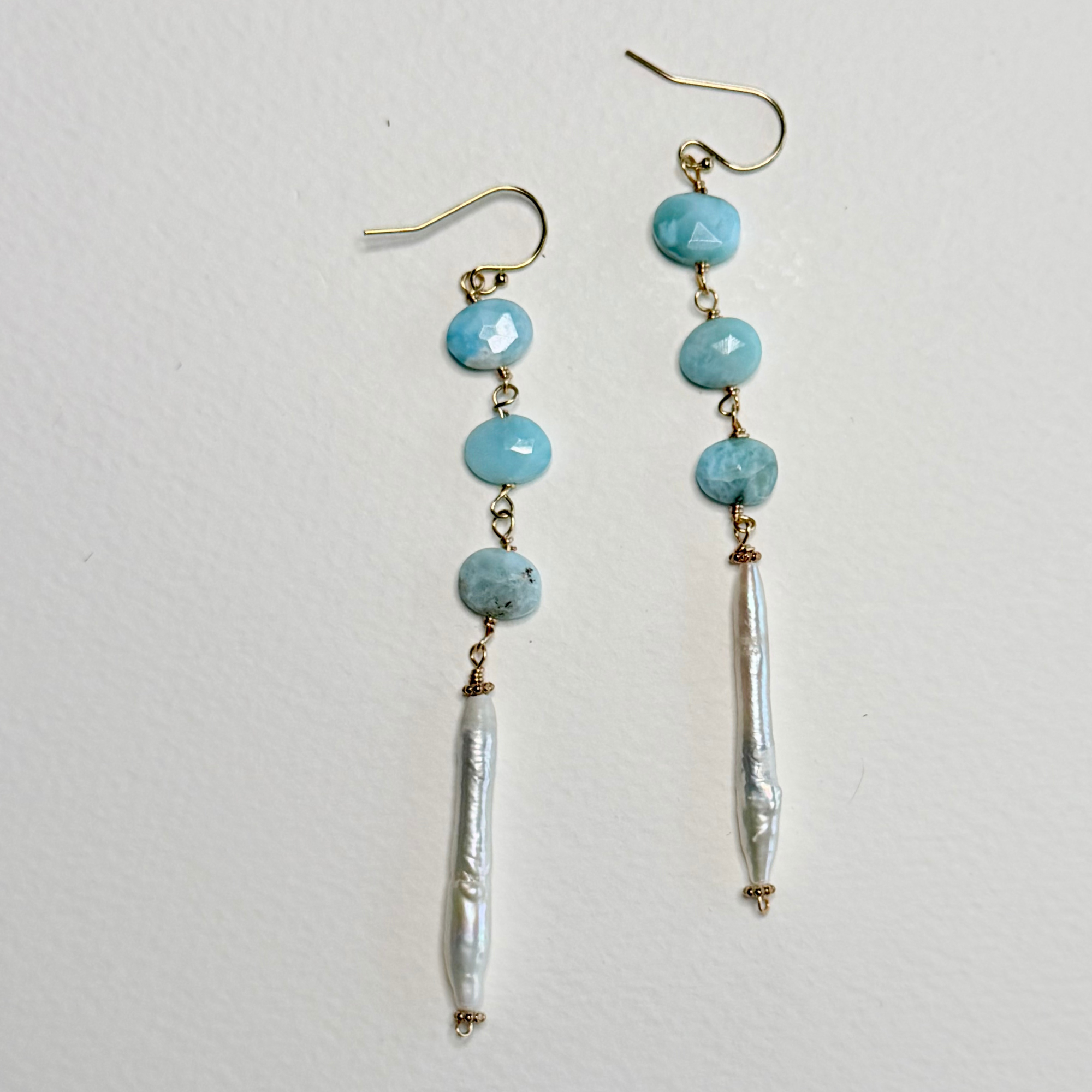 A pair of dangle earrings with light blue larimar faceted beads, gold-tone hooks and connectors, and a dangling baroque pearl bead, shown against a plain off-white background.