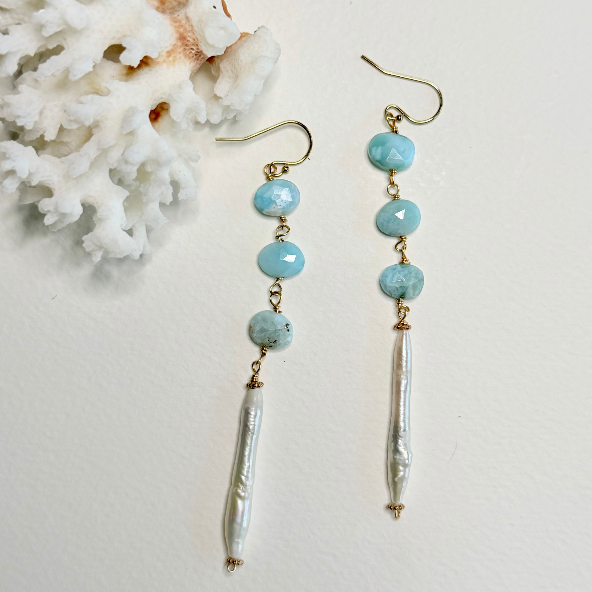 A pair of dangle earrings with light blue larimar faceted beads, gold-tone hooks, and white baroque pearl drops, displayed on a white textured surface next to a white coral shell.