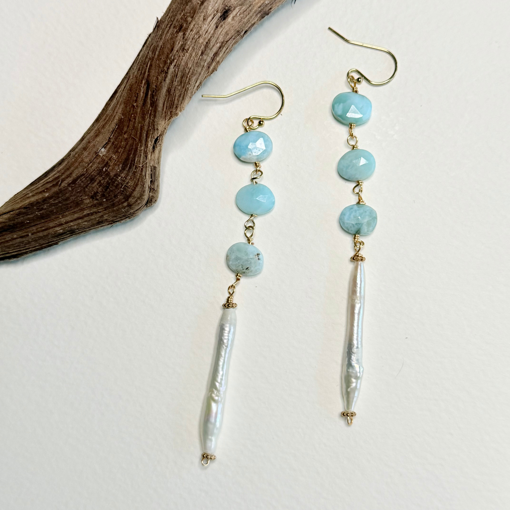 A pair of dangle earrings with light blue larimar faceted beads, gold-tone hooks, and white baroque pearl drops, displayed on a white background next to a piece of driftwood.