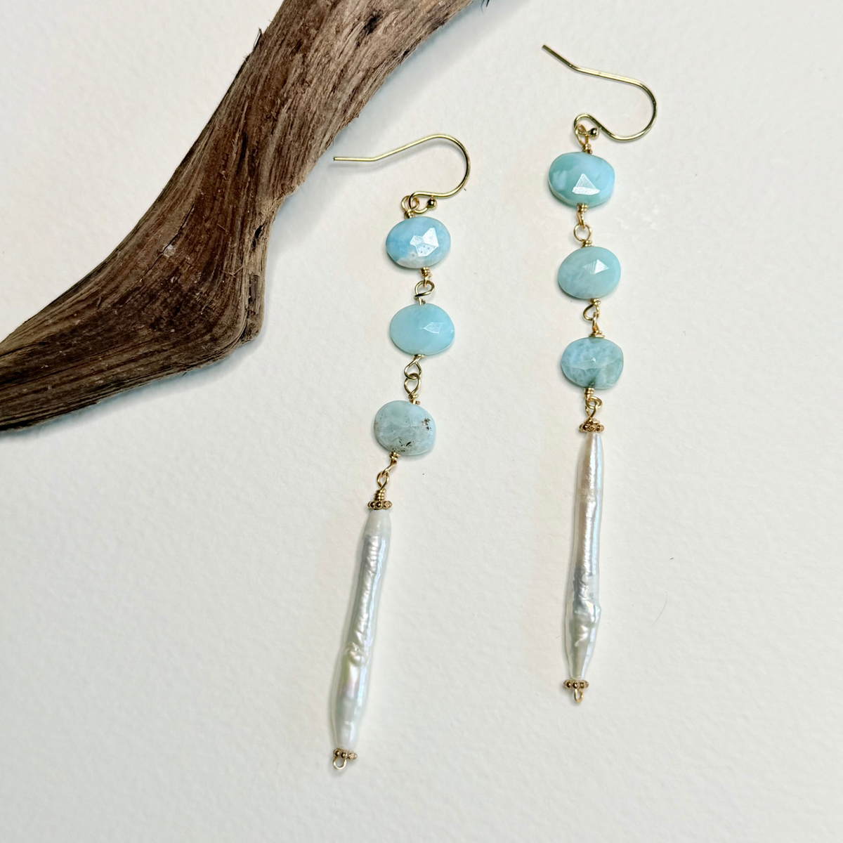 A pair of dangle earrings with light blue larimar faceted beads, gold-tone hooks, and white baroque pearl drops, displayed on a white background next to a piece of driftwood.