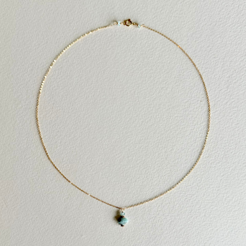 A delicate gold chain necklace with a small, dual-colored bead pendant featuring light blue and dark brown, displayed on a textured off-white background.