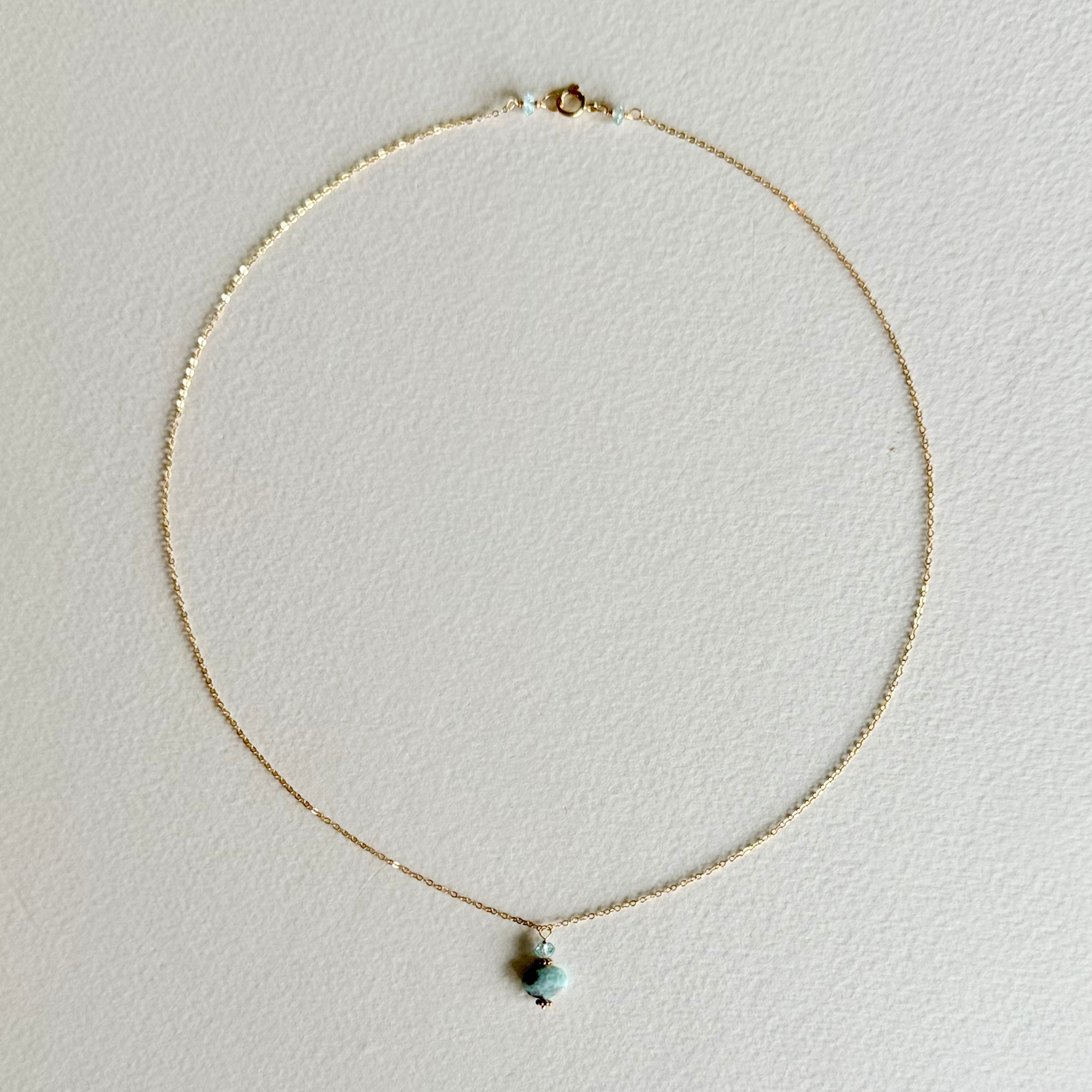 A delicate gold chain necklace with a small, dual-colored bead pendant featuring light blue and dark brown, displayed on a textured off-white background.