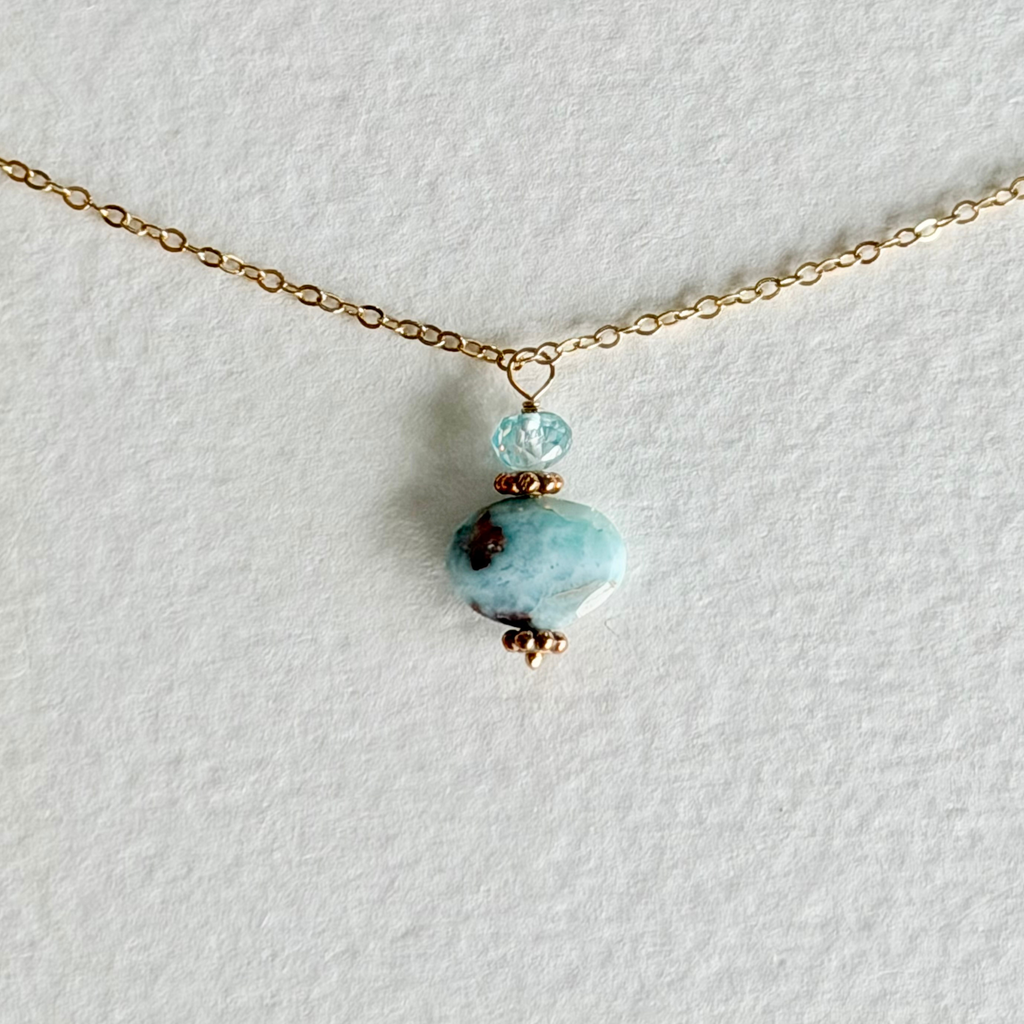 A delicate gold chain necklace featuring a dangle pendant with a small light blue zircon faceted bead, a larger larimar marbled light blue and white stone, and tiny gold beads, displayed on a textured white background.