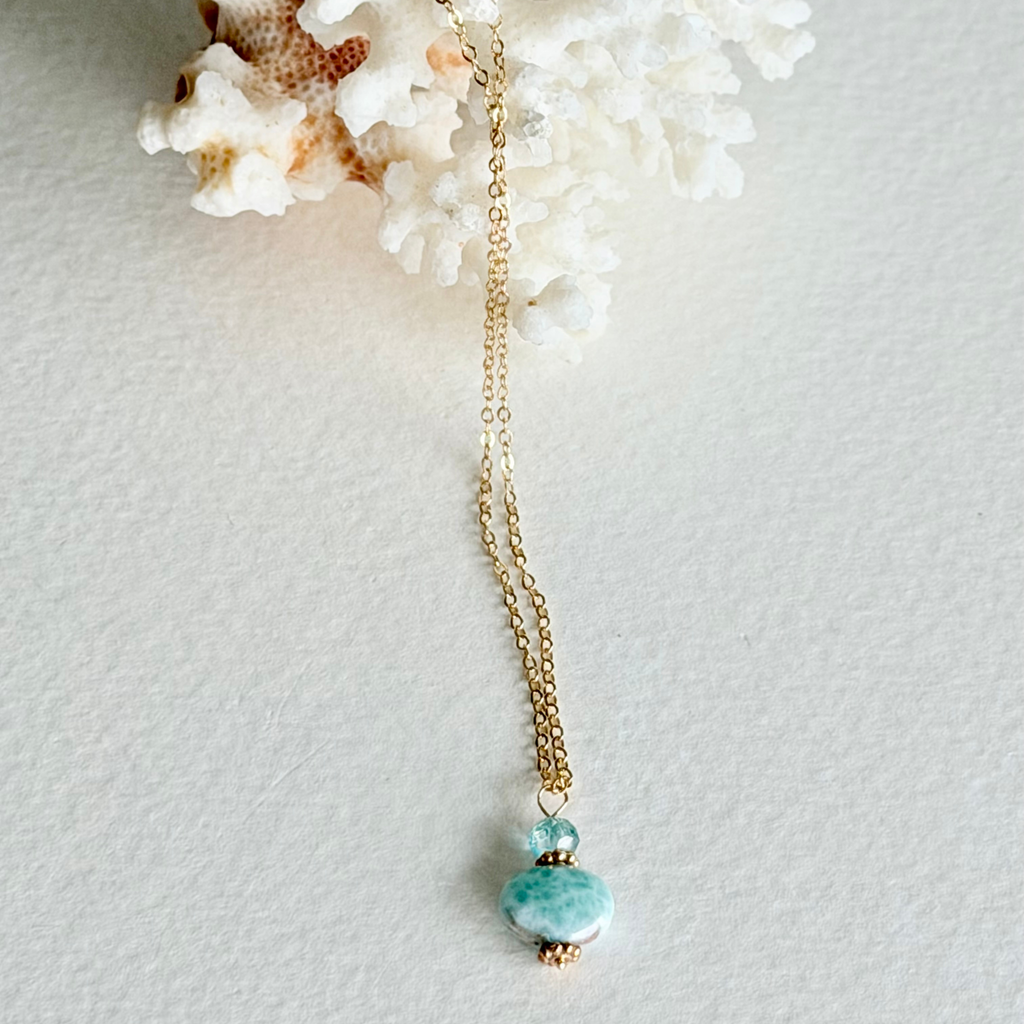 A gold chain necklace with a  pendant featuring a small zircon bead and a larger polished larimar stone, displayed on a textured white surface with white coral.