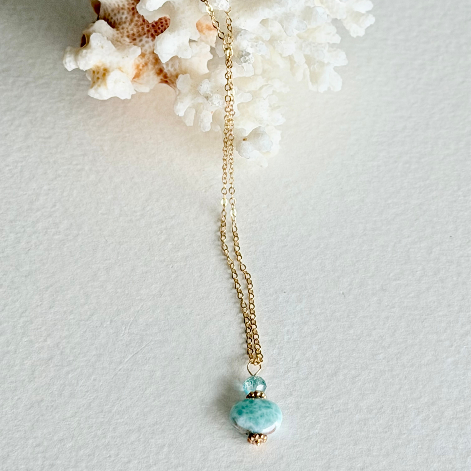 A gold chain necklace with a  pendant featuring a small zircon bead and a larger polished larimar stone, displayed on a textured white surface with white coral.