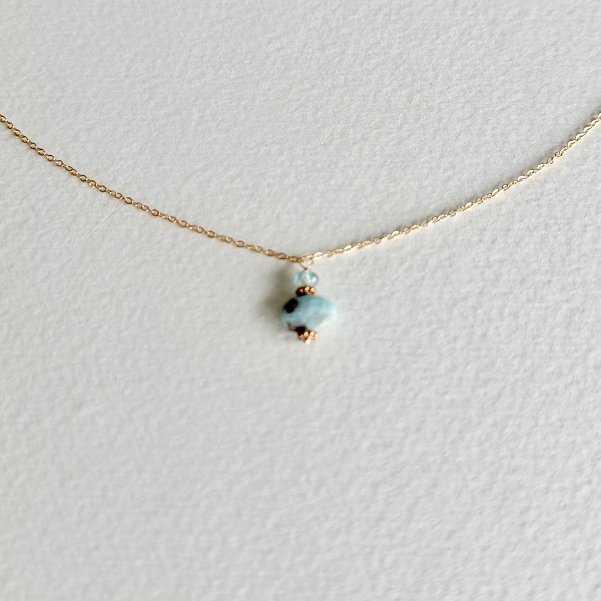 A delicate gold chain necklace featuring a small pendant with a light blue stone and tiny dark accents, displayed on a textured white background.