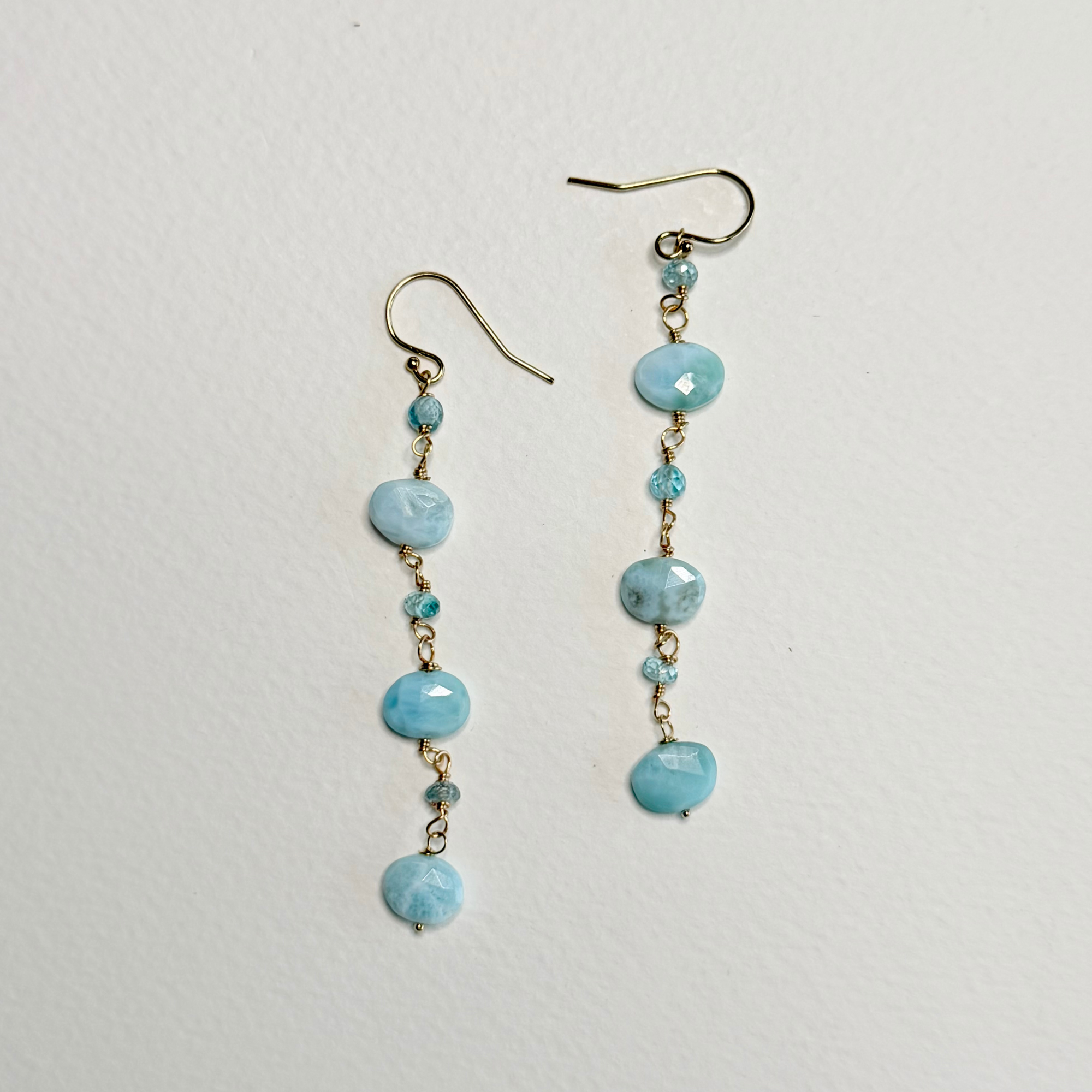 A pair of dangle earrings with light blue marbled beads and small faceted blue beads suspended from gold-tone fishhook wires, displayed on a textured off-white background.