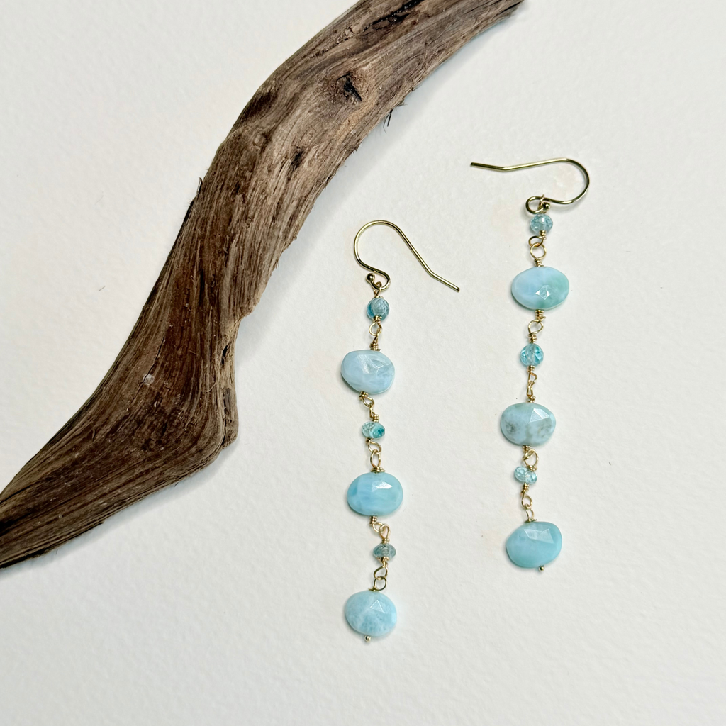 A pair of dangle earrings with light blue marbled beads and small gold-toned accents, displayed on a white background next to a piece of driftwood.
