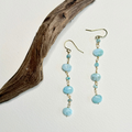 A pair of dangle earrings with light blue marbled beads and small gold-toned accents, displayed on a white background next to a piece of driftwood.