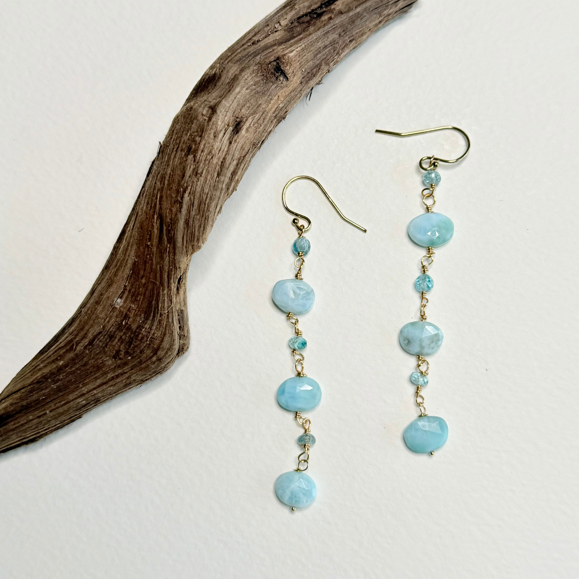 A pair of dangle earrings with light blue marbled beads and small gold-toned accents, displayed on a white background next to a piece of driftwood.