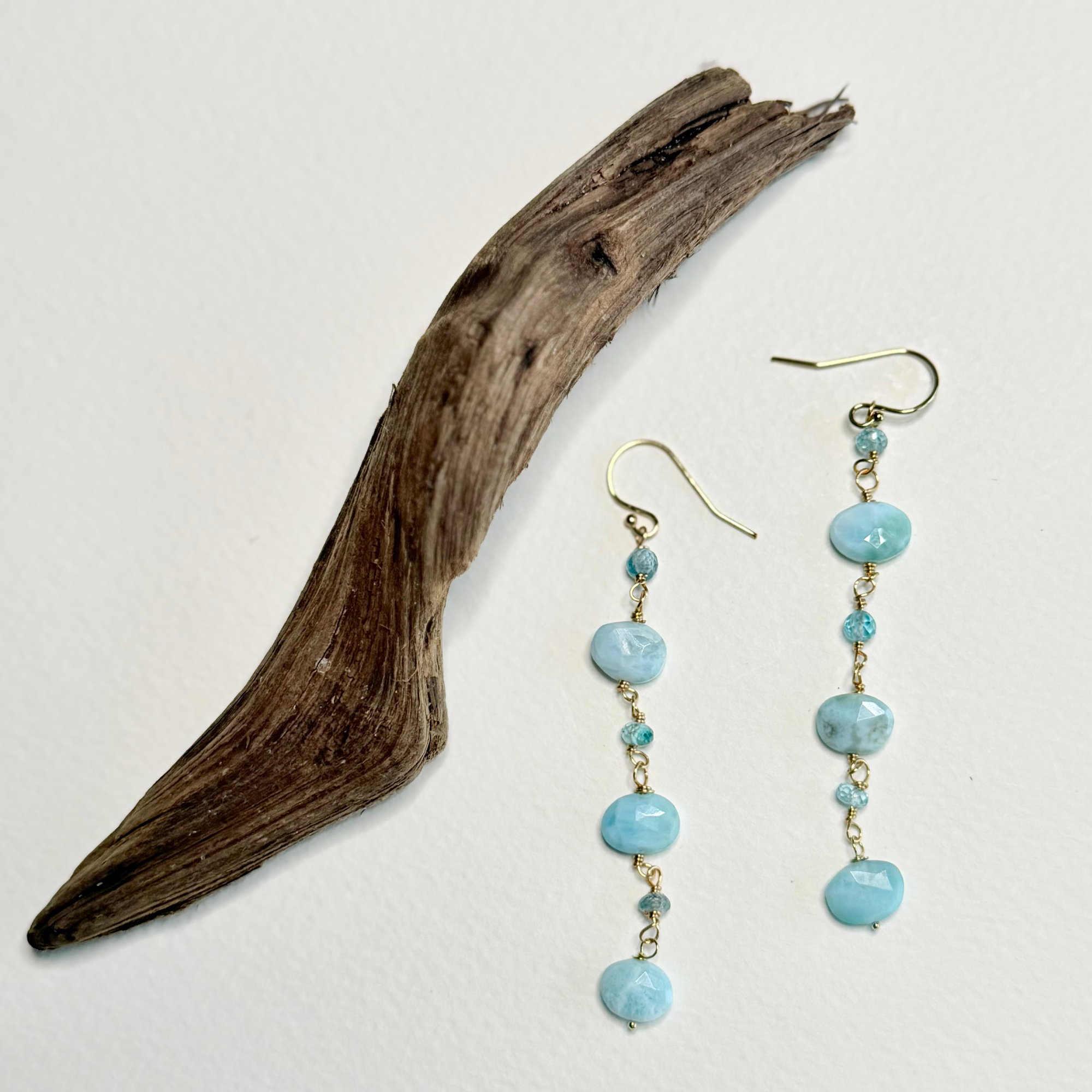 A pair of dangling earrings with light blue marbled beads and small gold-toned spacer beads, displayed beside a piece of driftwood on a plain white background.