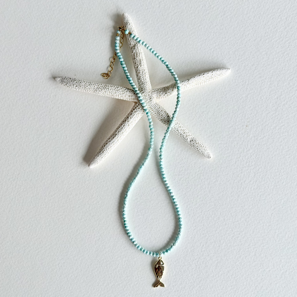 A beaded necklace with light aqua blue beads and a small gold mermaid tail charm, displayed on a white starfish prop against a white background.
