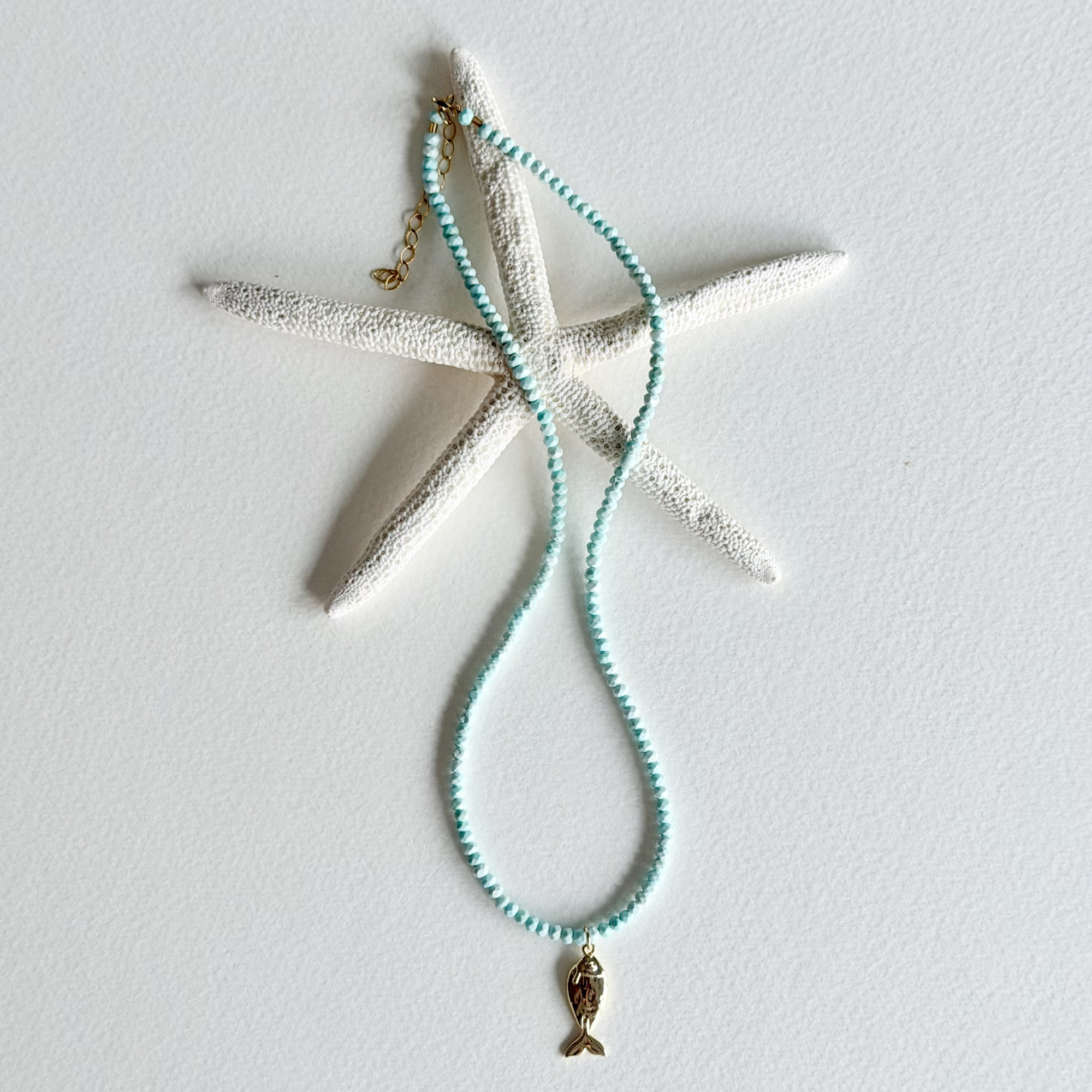 A beaded necklace with light aqua blue beads and a small gold mermaid tail charm, displayed on a white starfish prop against a white background.