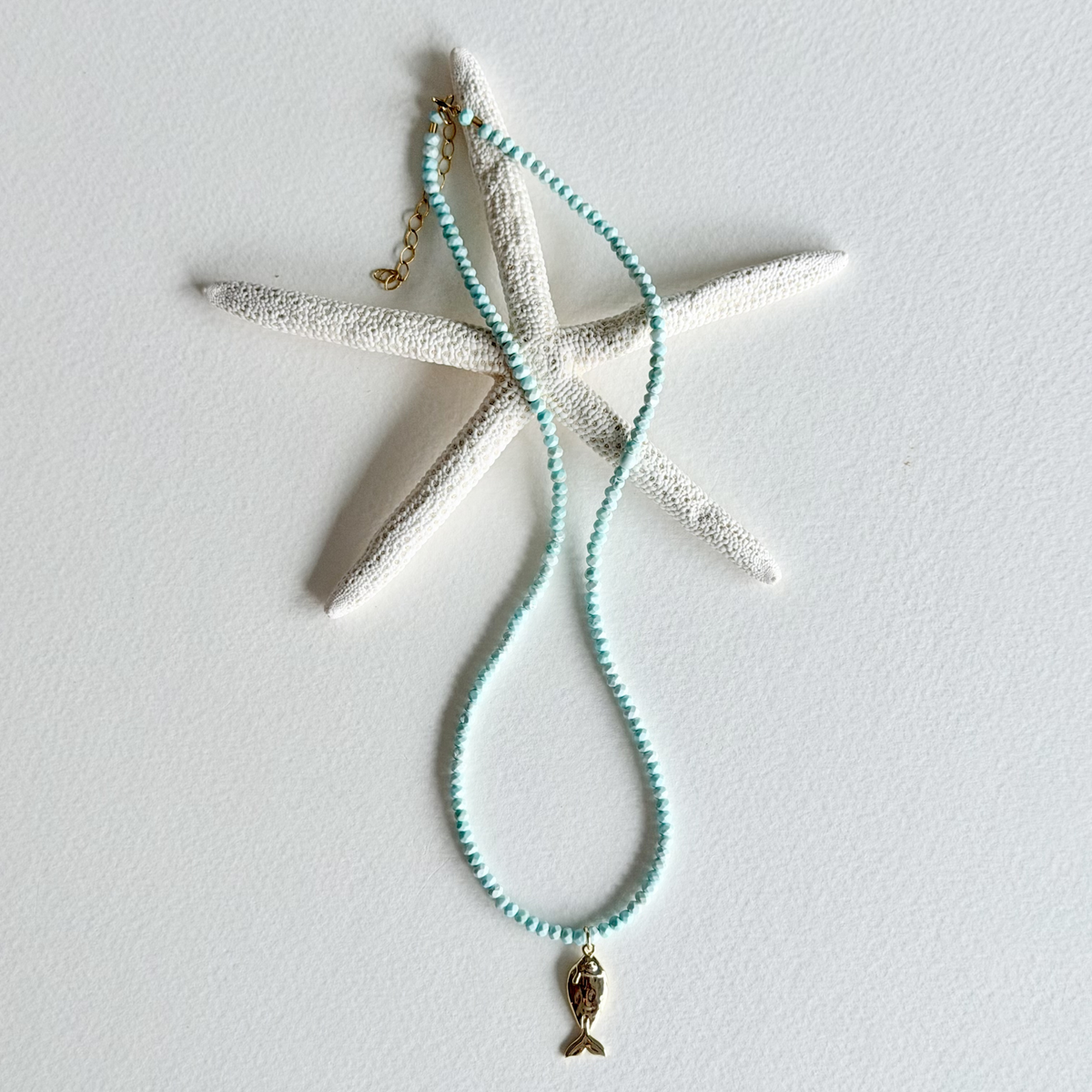 A beaded necklace with light aqua blue beads and a small gold mermaid tail charm, displayed on a white starfish prop against a white background.
