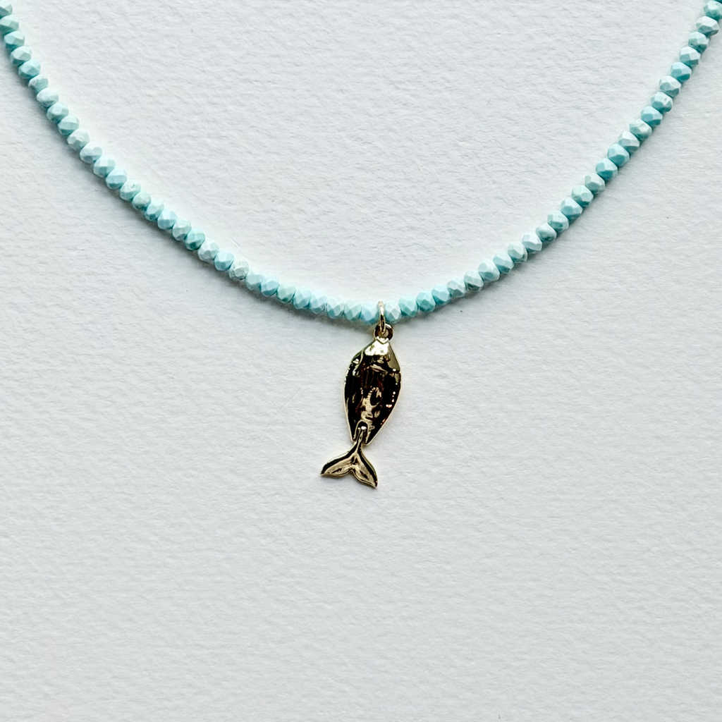 A delicate necklace featuring a strand of small, faceted light blue beads and a small gold fish charm, displayed on a textured white surface.