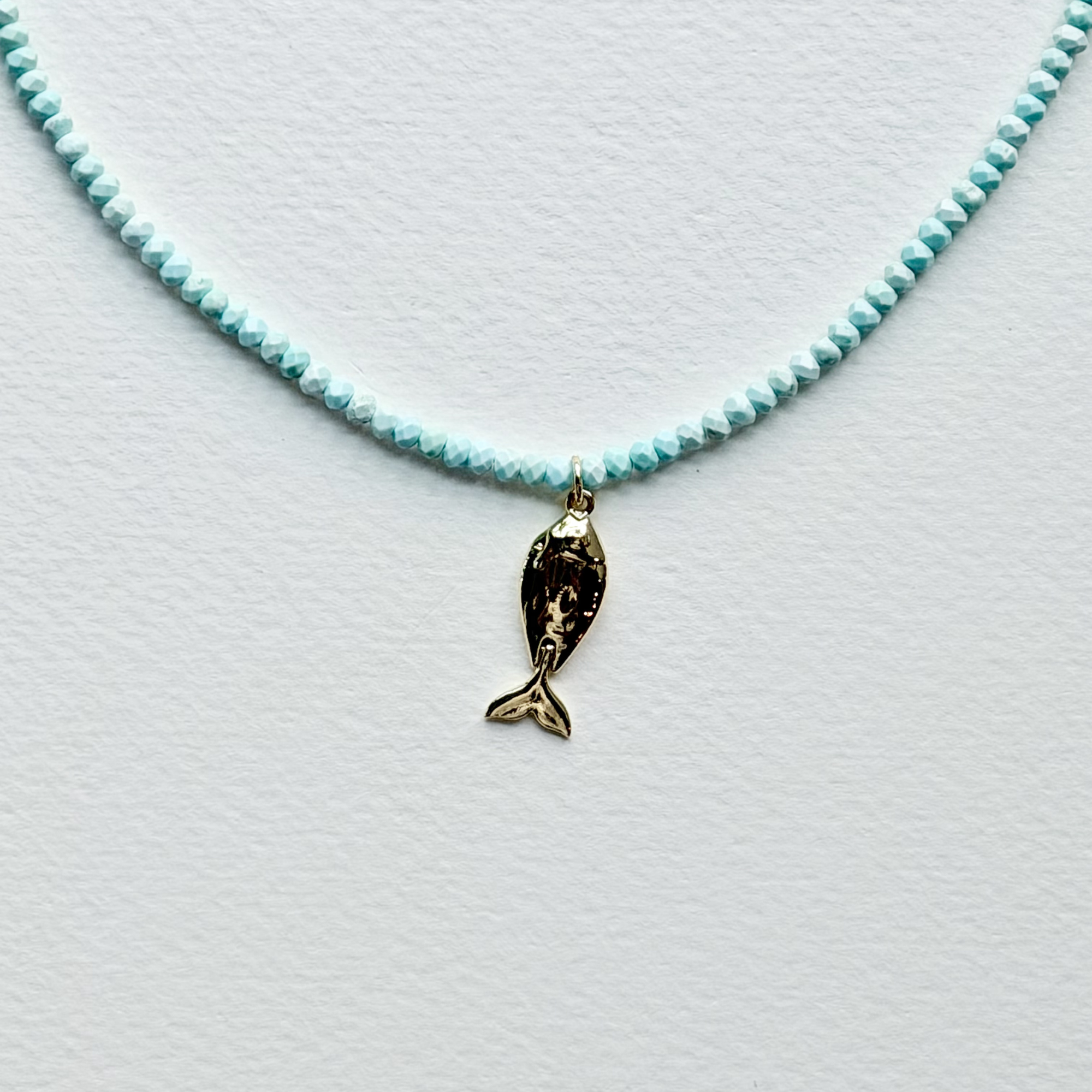 A delicate necklace featuring a strand of small, faceted light blue beads and a small gold fish charm, displayed on a textured white surface.