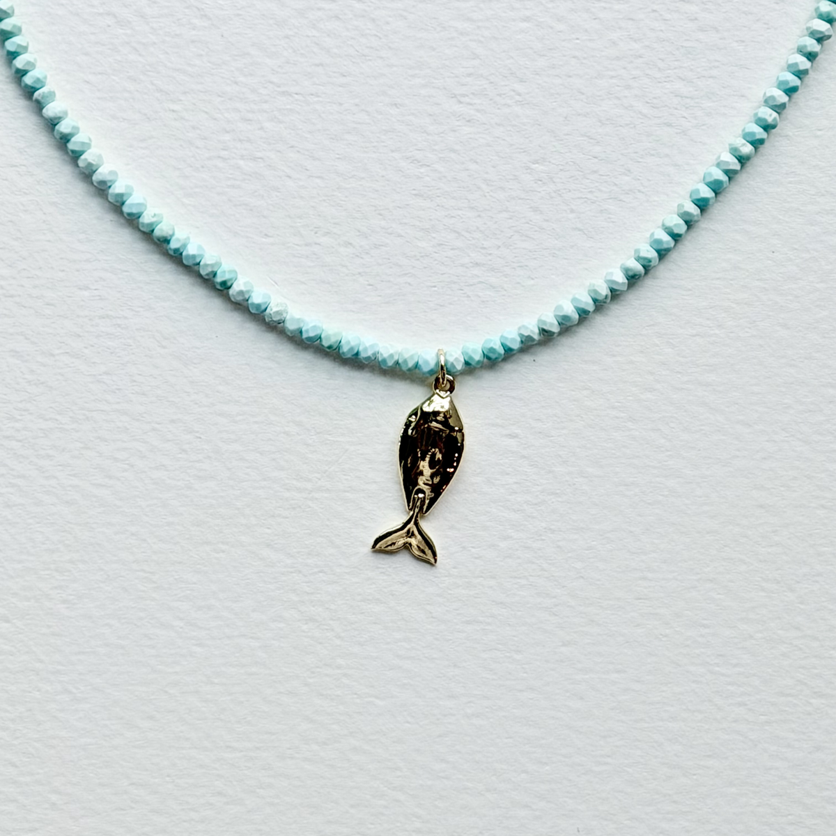 A delicate necklace featuring a strand of small, faceted light blue beads and a small gold fish charm, displayed on a textured white surface.
