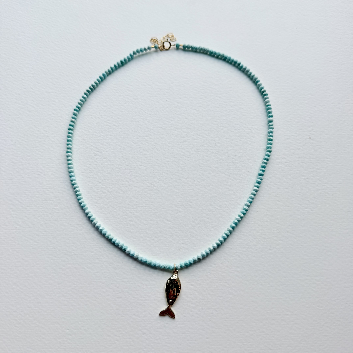 A delicate beaded necklace with small turquoise-blue beads and a dangling fish charm, displayed on a light gray textured background.