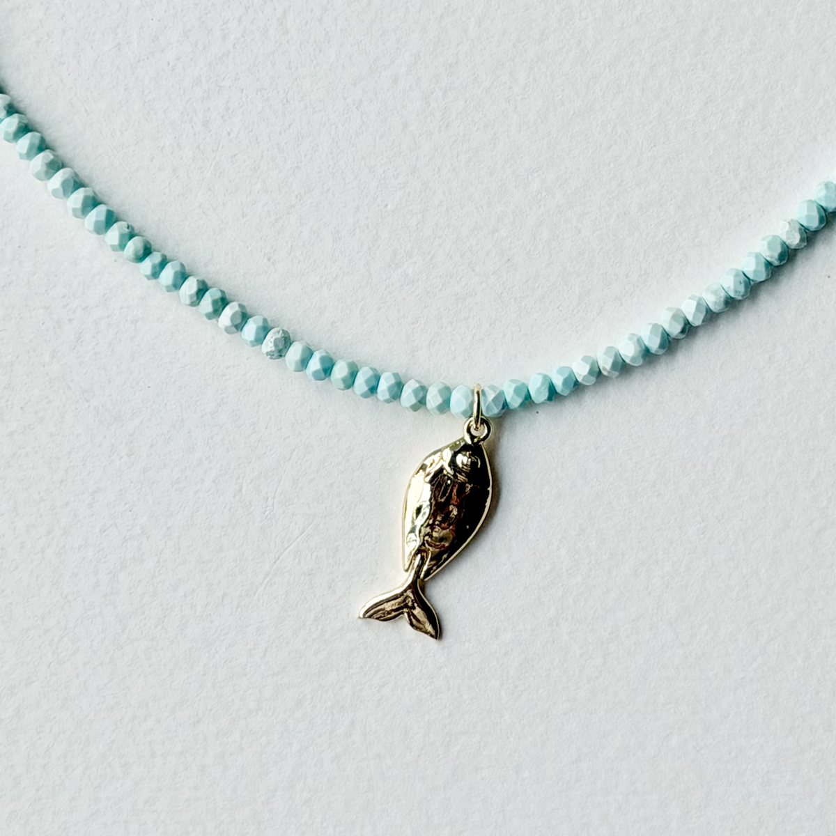 A delicate necklace featuring a strand of small, light blue faceted beads and a central gold fish-shaped charm, displayed on a textured white surface.