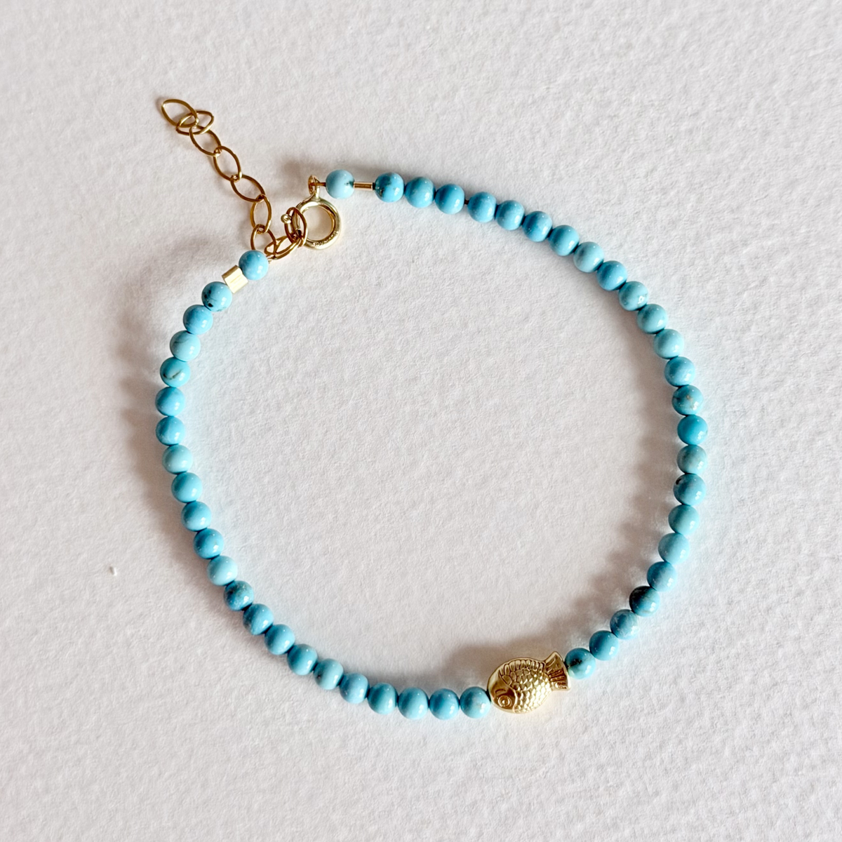 Turquoise beaded bracelet with a gold fish charm on a light gray background