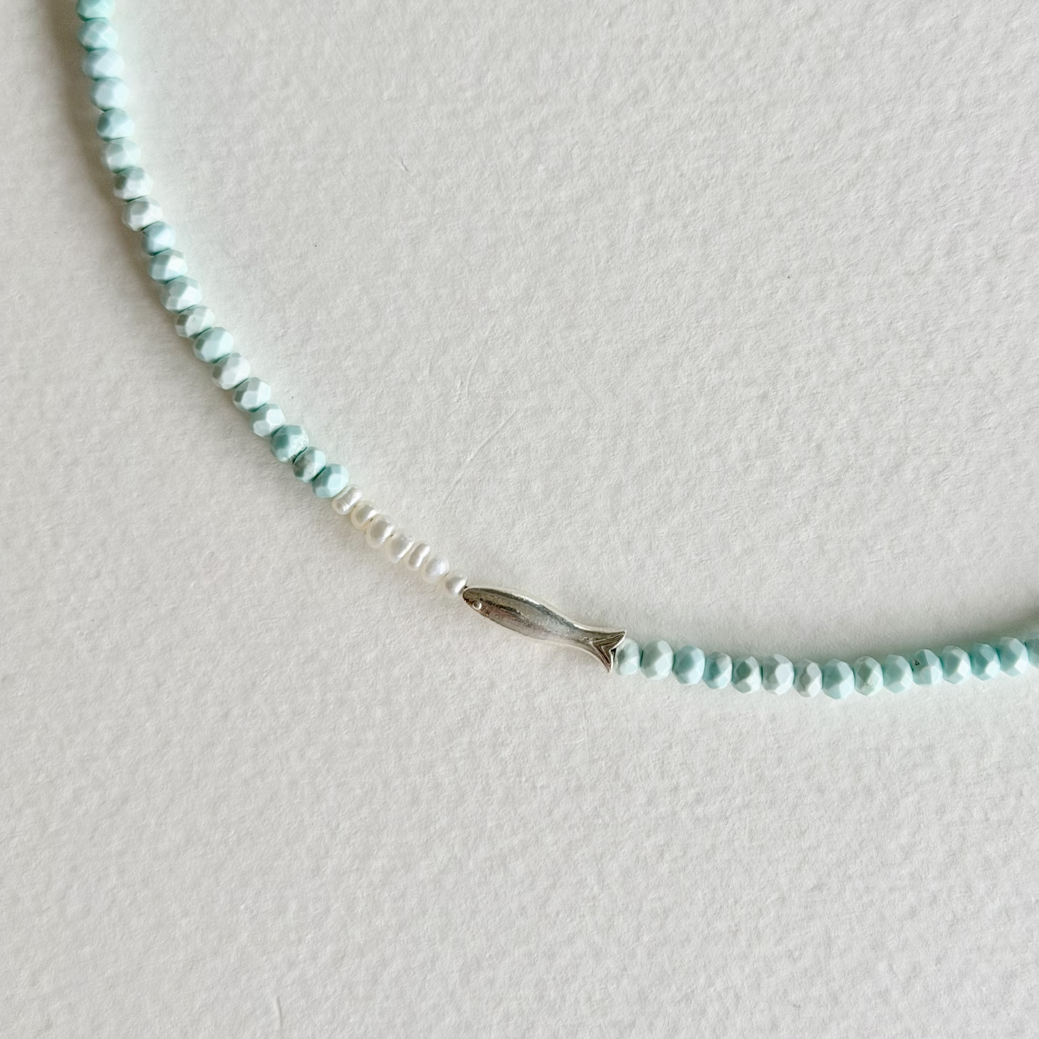 A delicate beaded necklace featuring light blue-green beads, a short section of small white beads, and a small silver fish charm, displayed on a textured off-white surface.