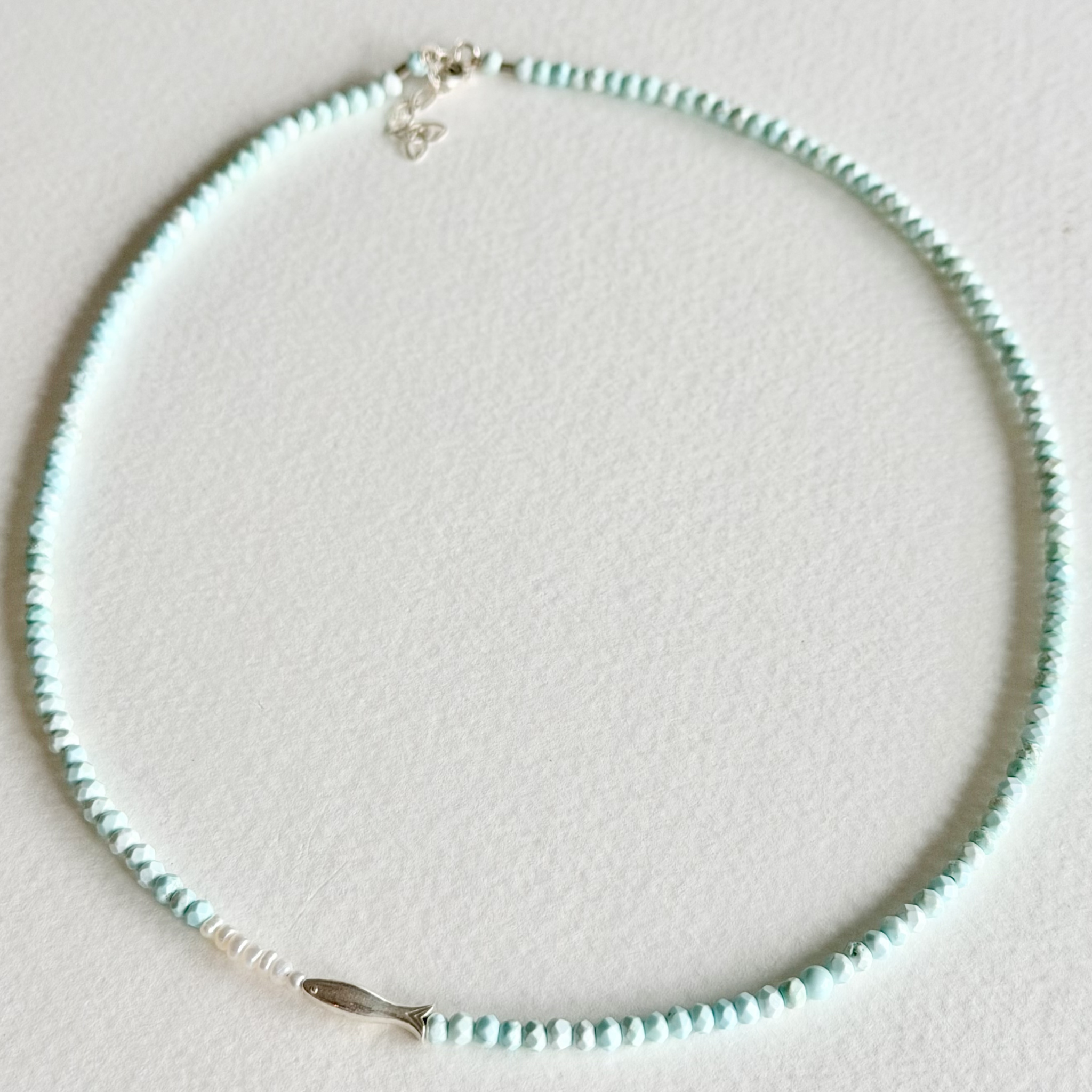 A delicate beaded necklace with light blue-green beads, a small silver fish charm, and a silver lobster clasp, displayed on a textured white surface.