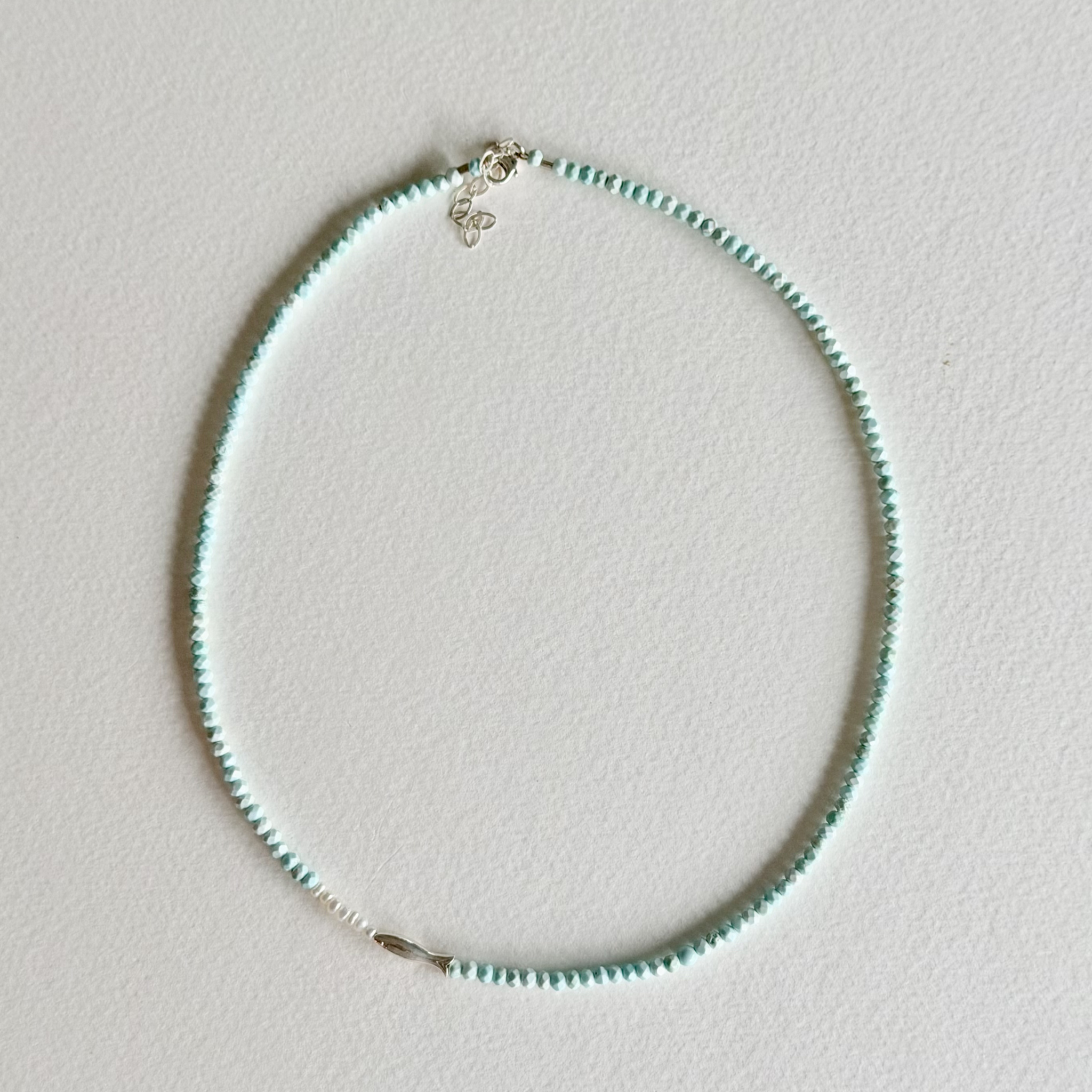 A delicate beaded necklace with small mint green beads and silver-toned hardware, laid flat on a light off-white textured surface.