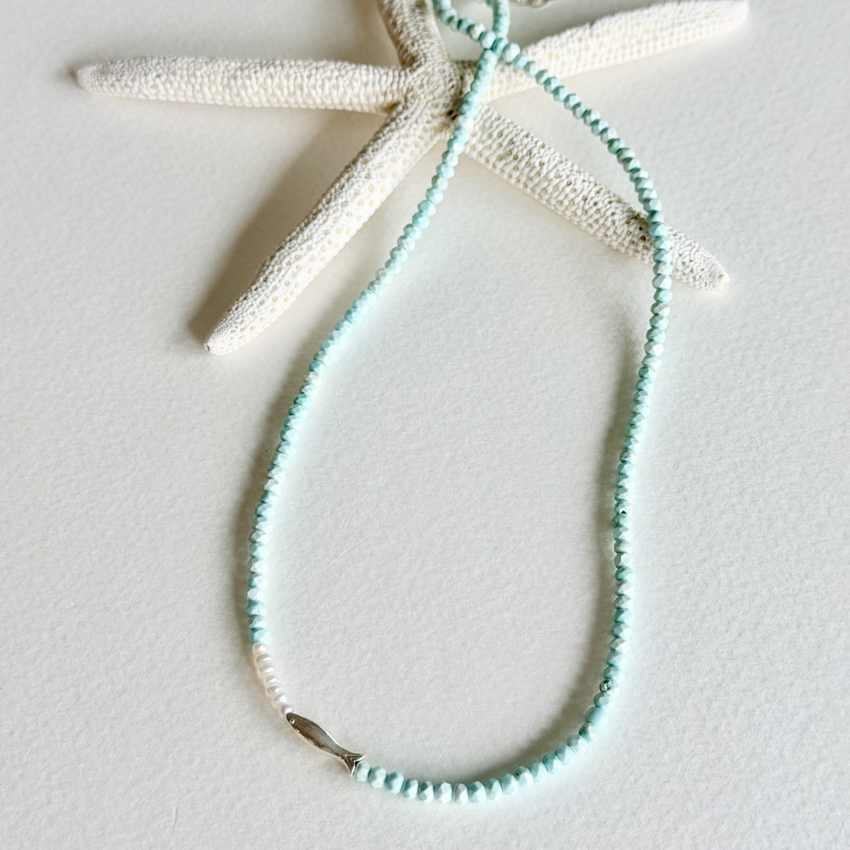 A delicate beaded necklace in light aqua blue, styled on a textured white background with a dried starfish prop.