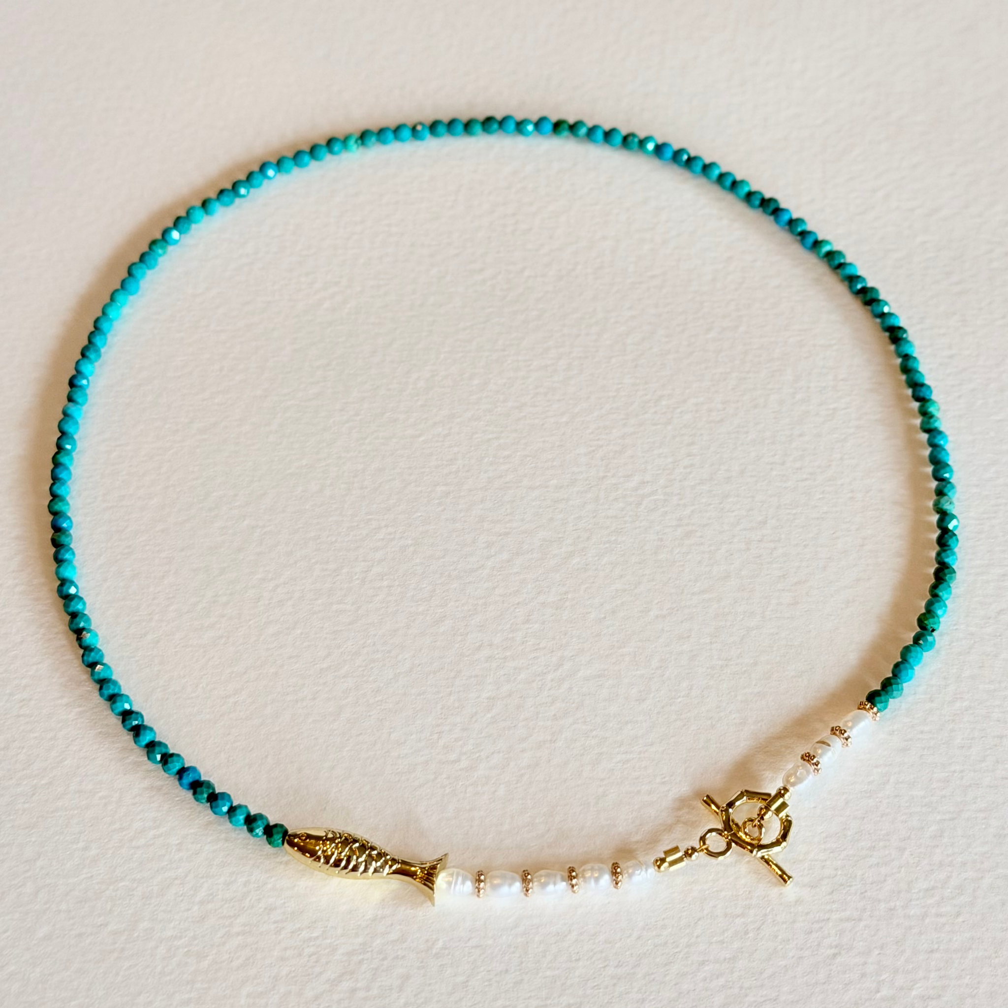 Turquoise beaded necklace with gold fish and anchor charms on a light background