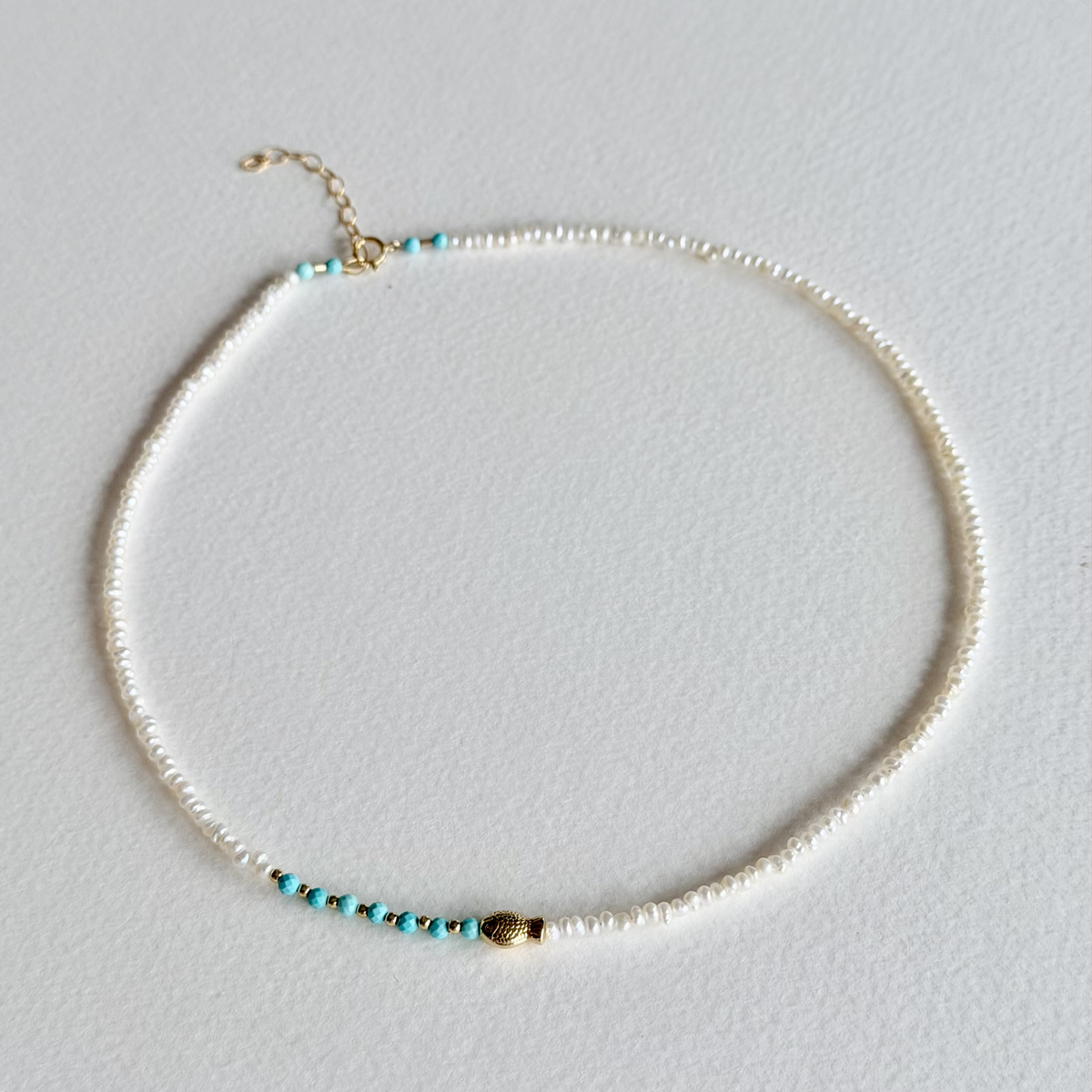 A delicate beaded necklace with small white pearls, turquoise blue beads, and gold-toned accents, displayed on a light gray textured background.