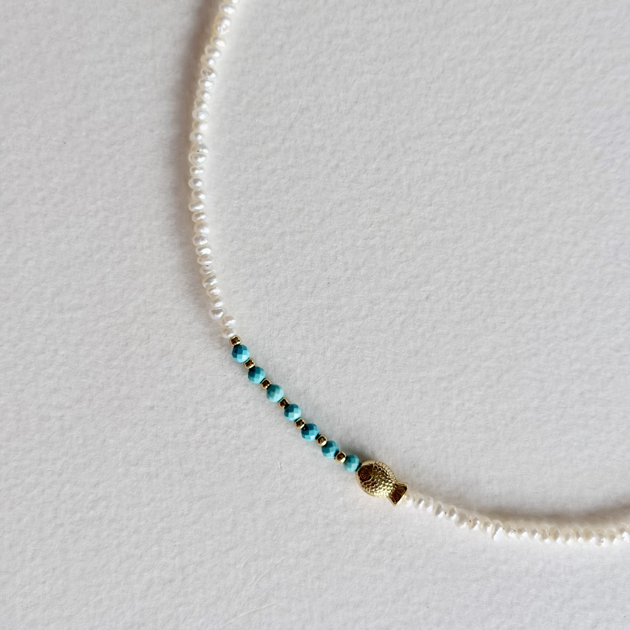 A delicate necklace featuring small white pearl-like beads with a central strand of turquoise blue beads separated by tiny gold spacers, finished with a textured gold bead accent.
