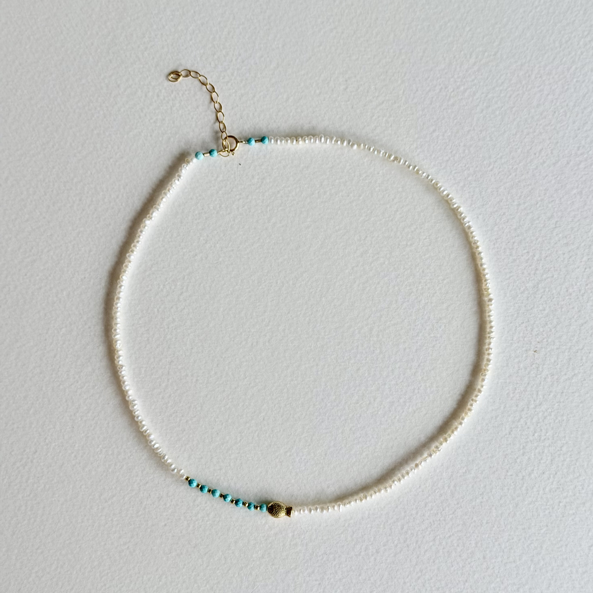 A delicate beaded necklace with small white beads, turquoise blue spacer beads, and gold-toned metal accents, including a small charm and extender chain, displayed on a light gray textured background.