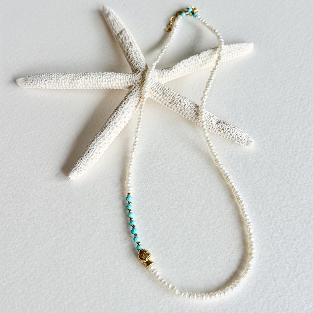 A delicate beaded necklace with white beads, a small turquoise bead accent, and gold-toned metal details, displayed on a white starfish prop against a white background.