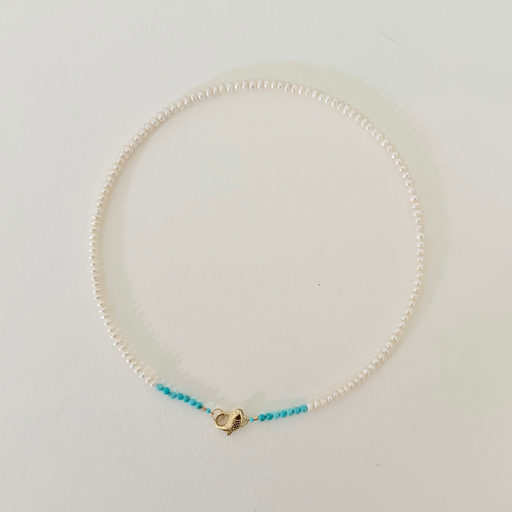 A delicate necklace made of small white pearls with a small section of turquoise blue beads on each side, connected by a gold heart-shaped clasp, displayed on a clean white background.