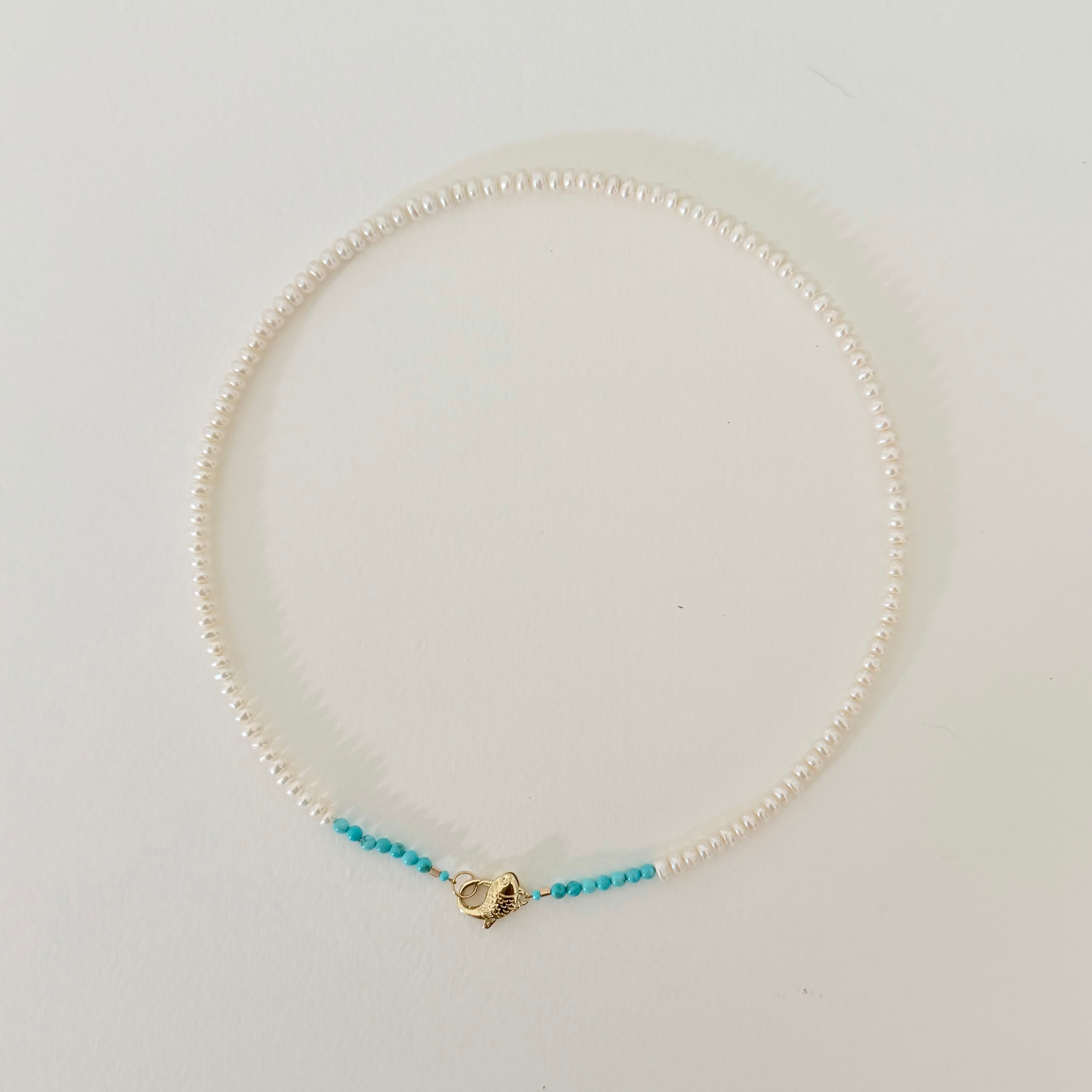 A delicate necklace made of small white pearls with a small section of turquoise blue beads on each side, connected by a gold heart-shaped clasp, displayed on a clean white background.