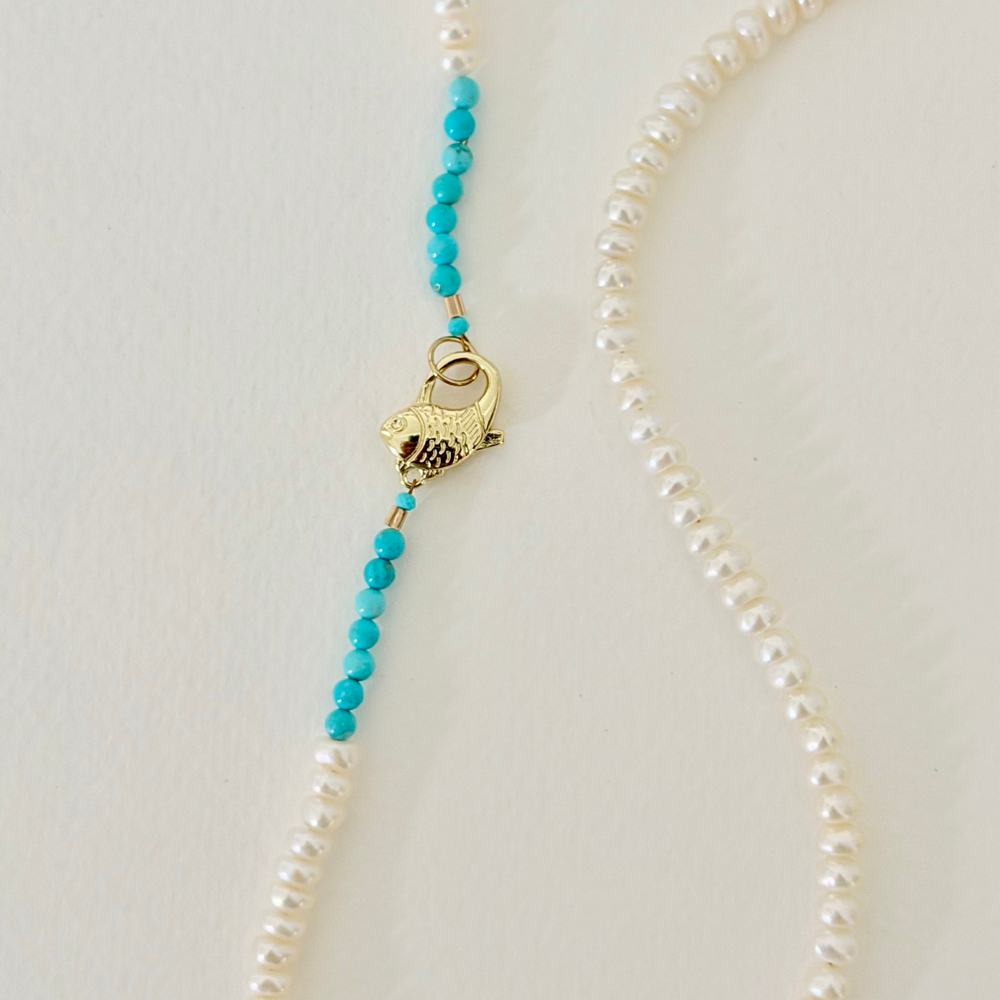 A pearl necklace with a decorative gold clasp featuring a small fish design, accented with bright turquoise beads on a light beige background.