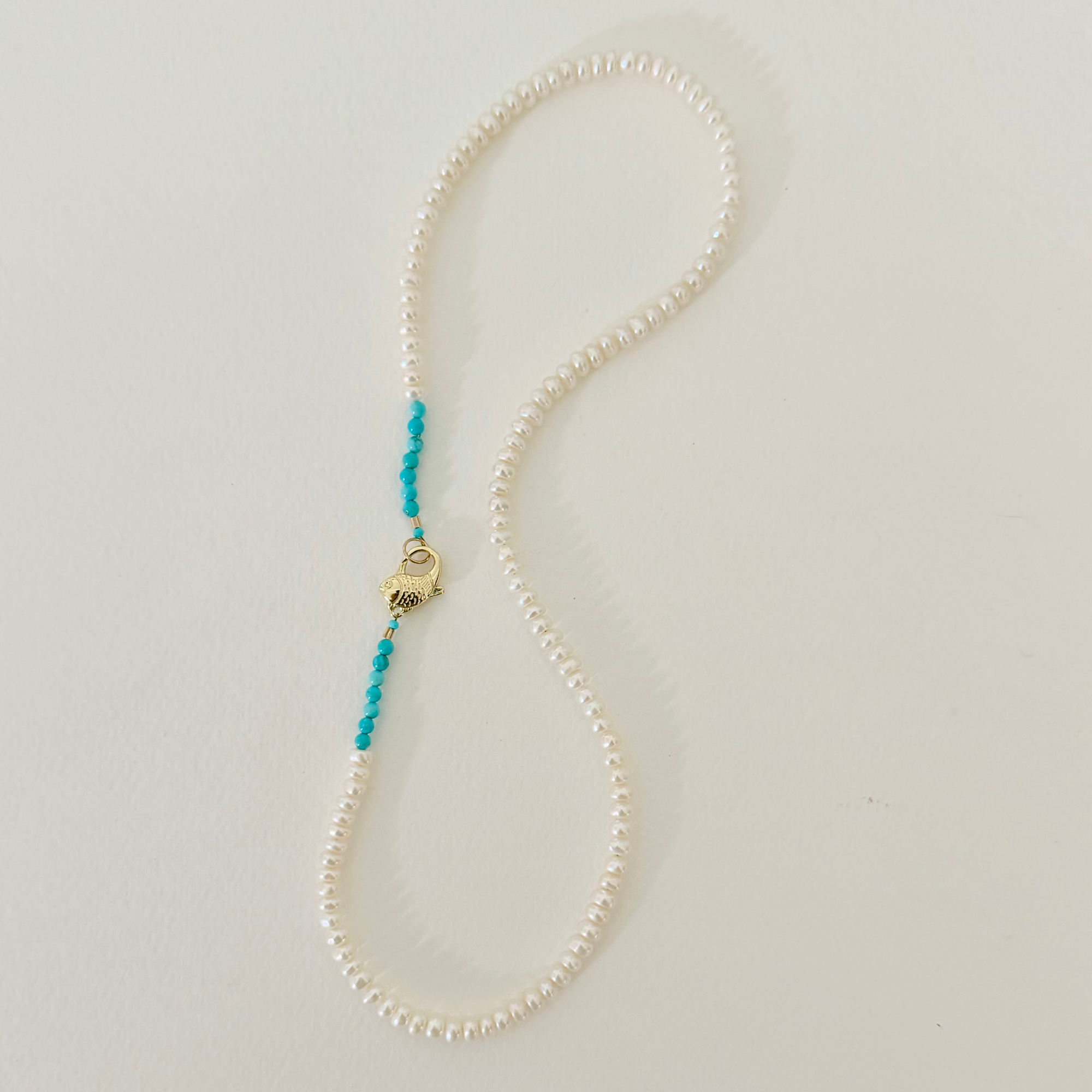 A white pearl necklace with a gold hamsa hand charm and turquoise blue bead accents, displayed on a light off-white background.