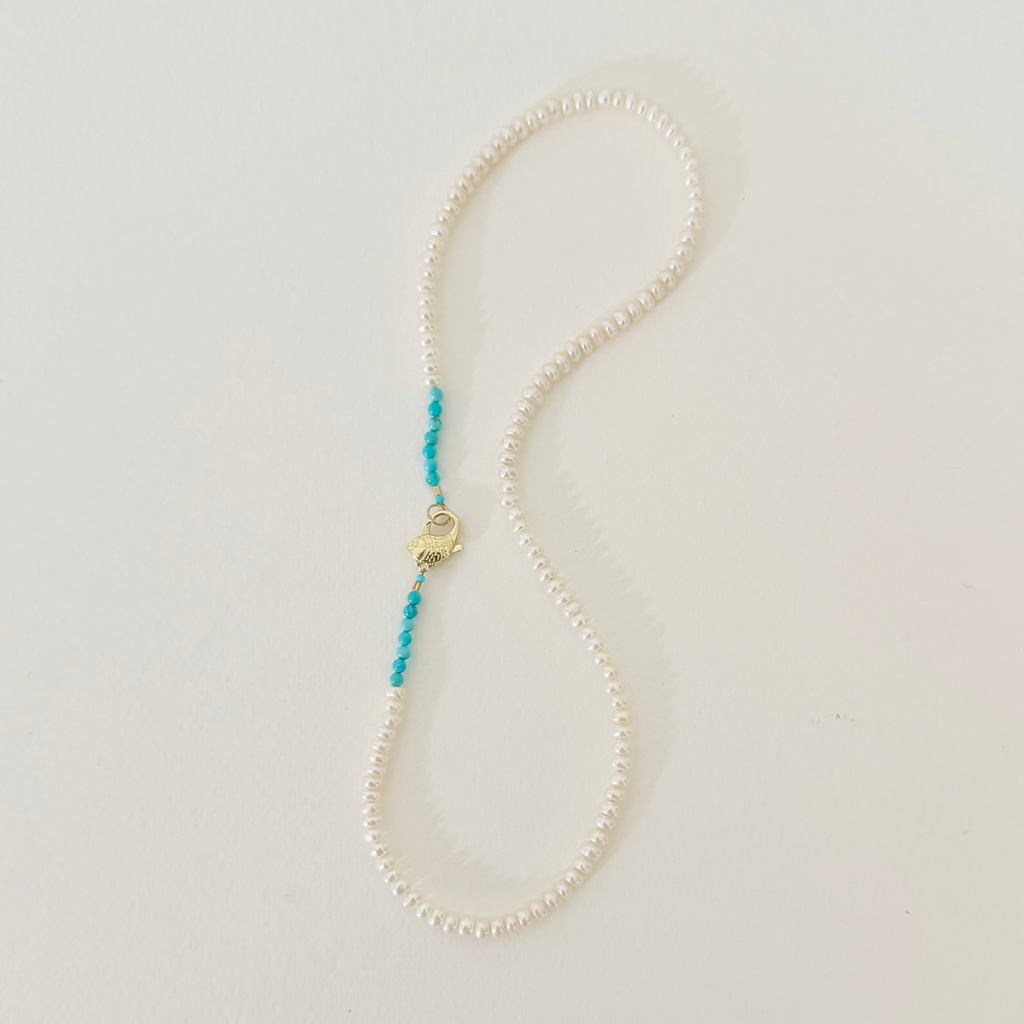 A delicate necklace made of small white pearls with a short turquoise beaded accent and a gold lobster clasp, displayed on a light off-white background.