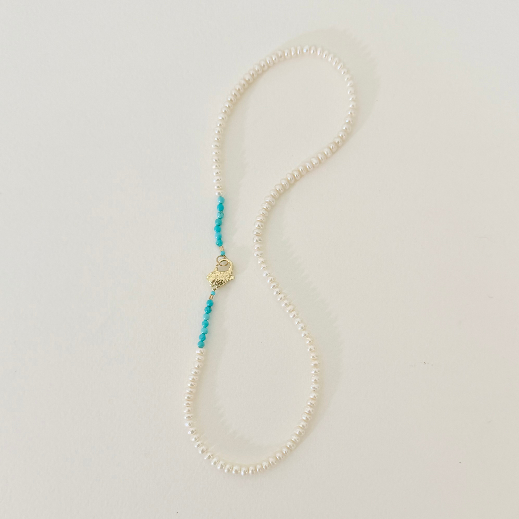 A delicate necklace made of small white pearls with a short turquoise beaded accent and a gold lobster clasp, displayed on a light off-white background.