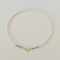 A delicate pearl necklace with a small gold heart-shaped clasp and two strands of tiny turquoise blue beads, displayed on a light cream background.