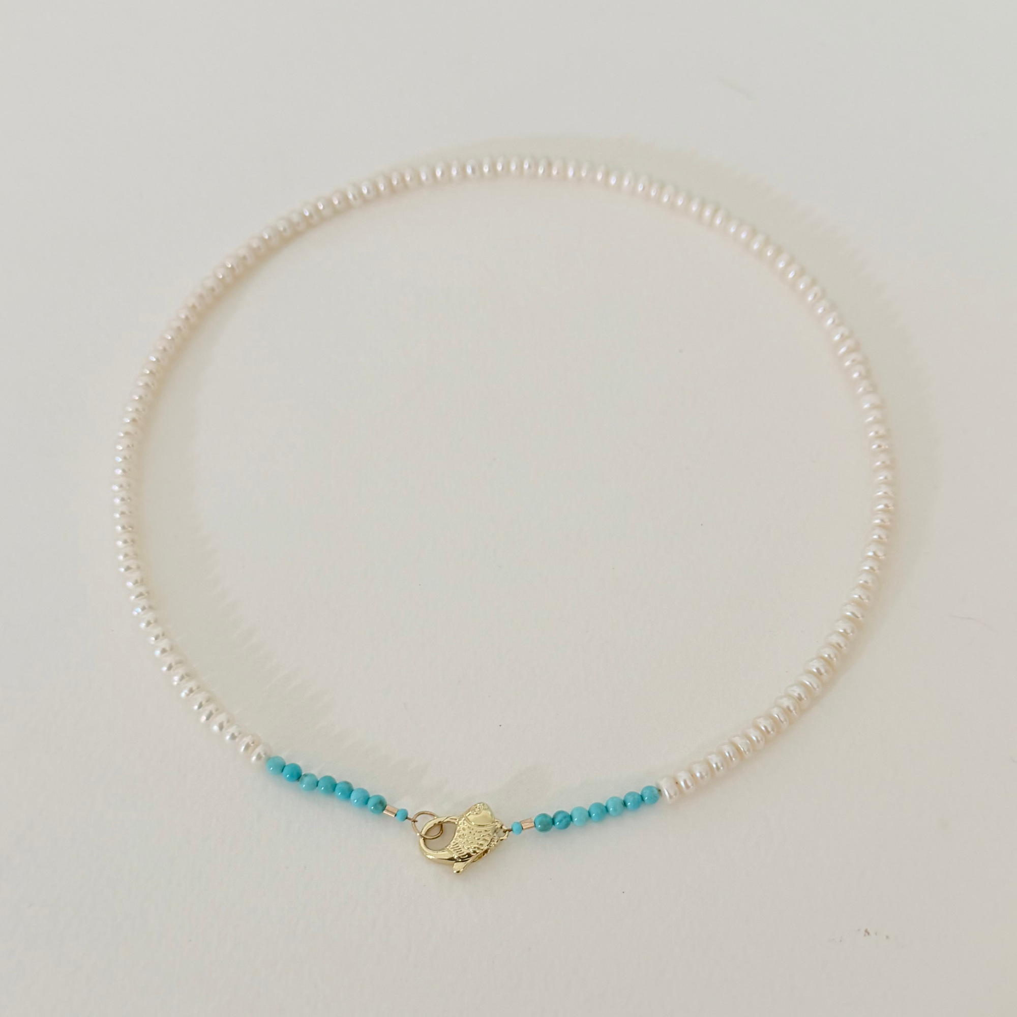 A delicate pearl necklace with a small gold heart-shaped clasp and two strands of tiny turquoise blue beads, displayed on a light cream background.