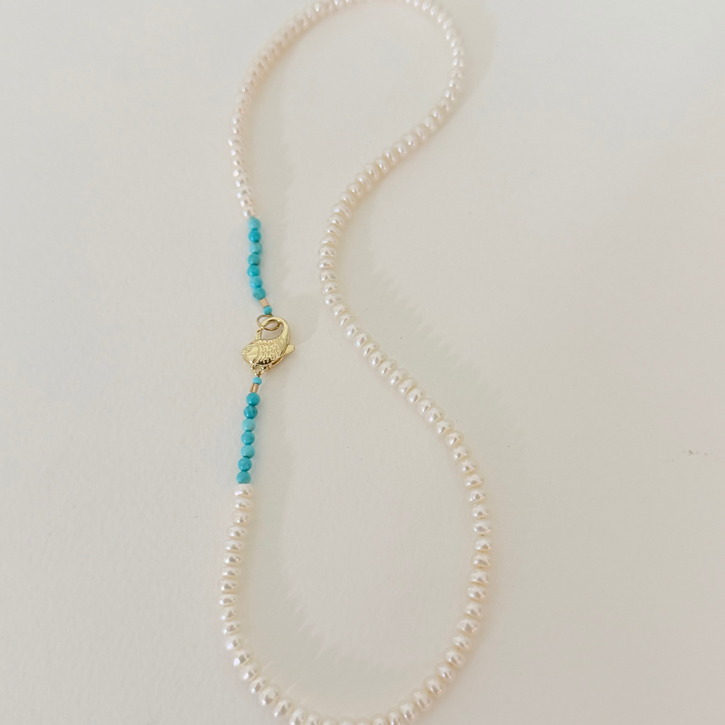 A delicate pearl necklace with a small gold heart-shaped charm and two strands of turquoise blue beads, displayed on a light beige background.