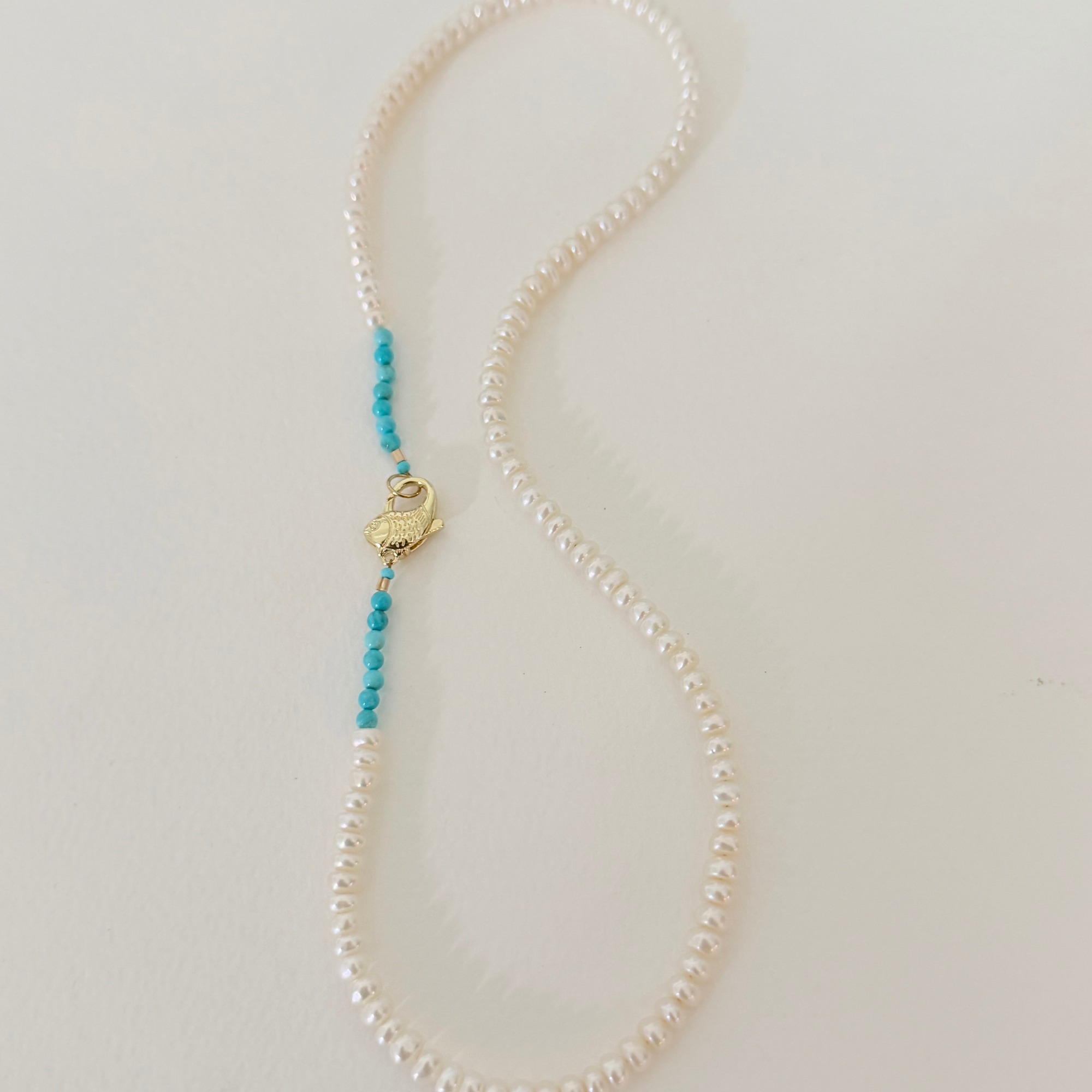 A delicate pearl necklace with a small gold heart-shaped charm and two strands of turquoise blue beads, displayed on a light beige background.
