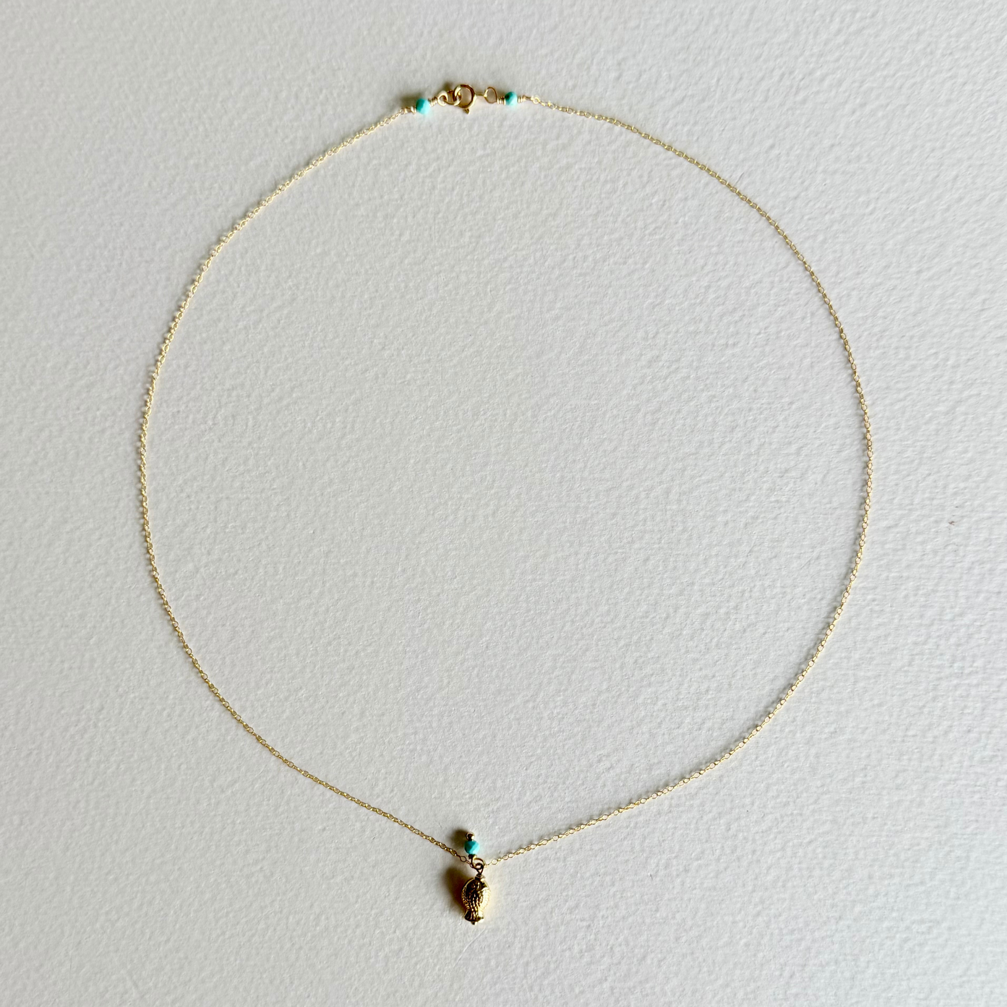 A delicate gold chain necklace with a small gold pendant and tiny turquoise accent beads, displayed on a textured off-white background.