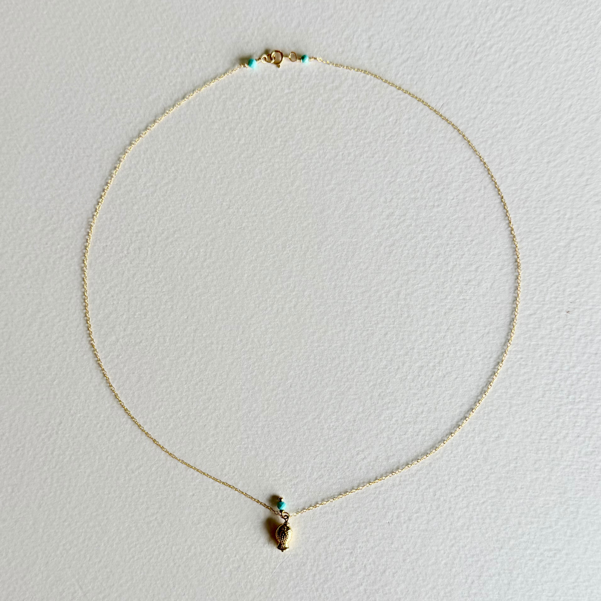 A delicate gold chain necklace with a small gold pendant and tiny turquoise accent beads, displayed on a textured off-white background.