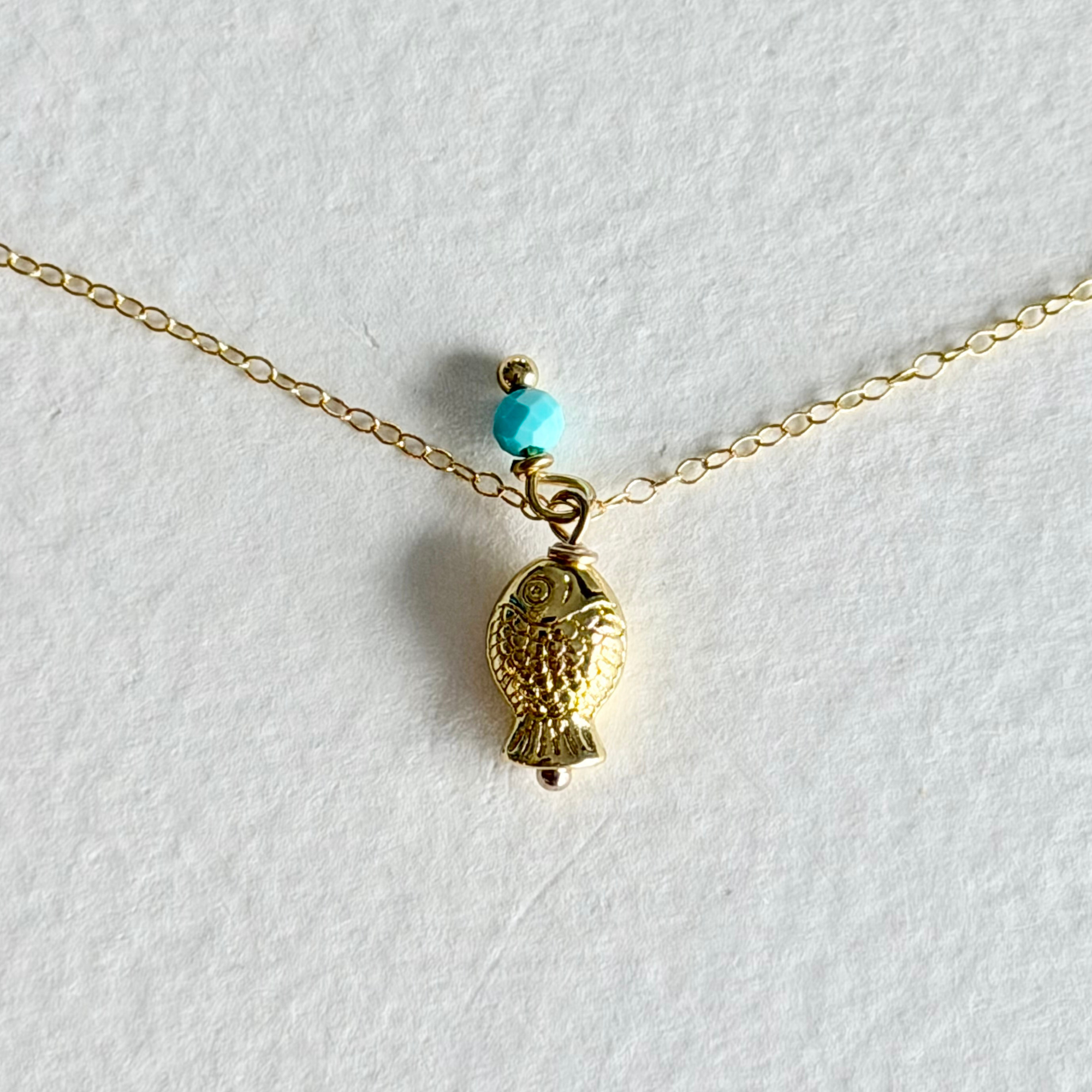 A delicate gold chain necklace featuring a small fish-shaped pendant and a single turquoise bead, displayed on a textured white background.