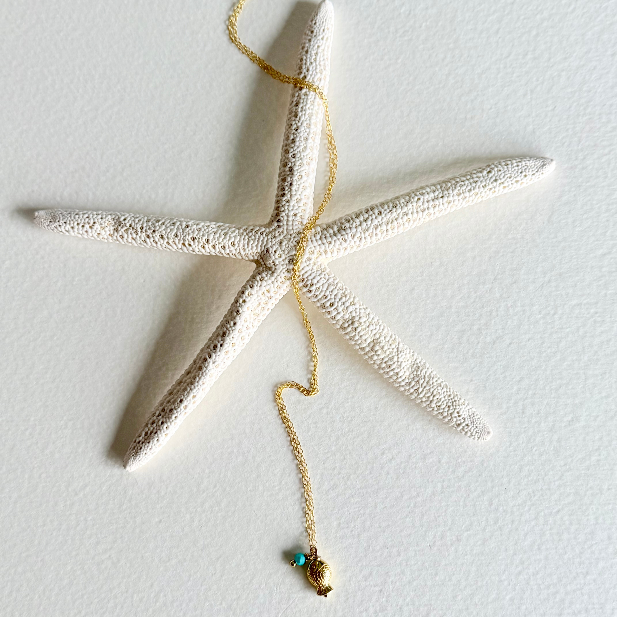 A textured white starfish wall hanging with a gold-toned chain draped over it and a small shell and turquoise pendant at the end, shown against a plain white wall.