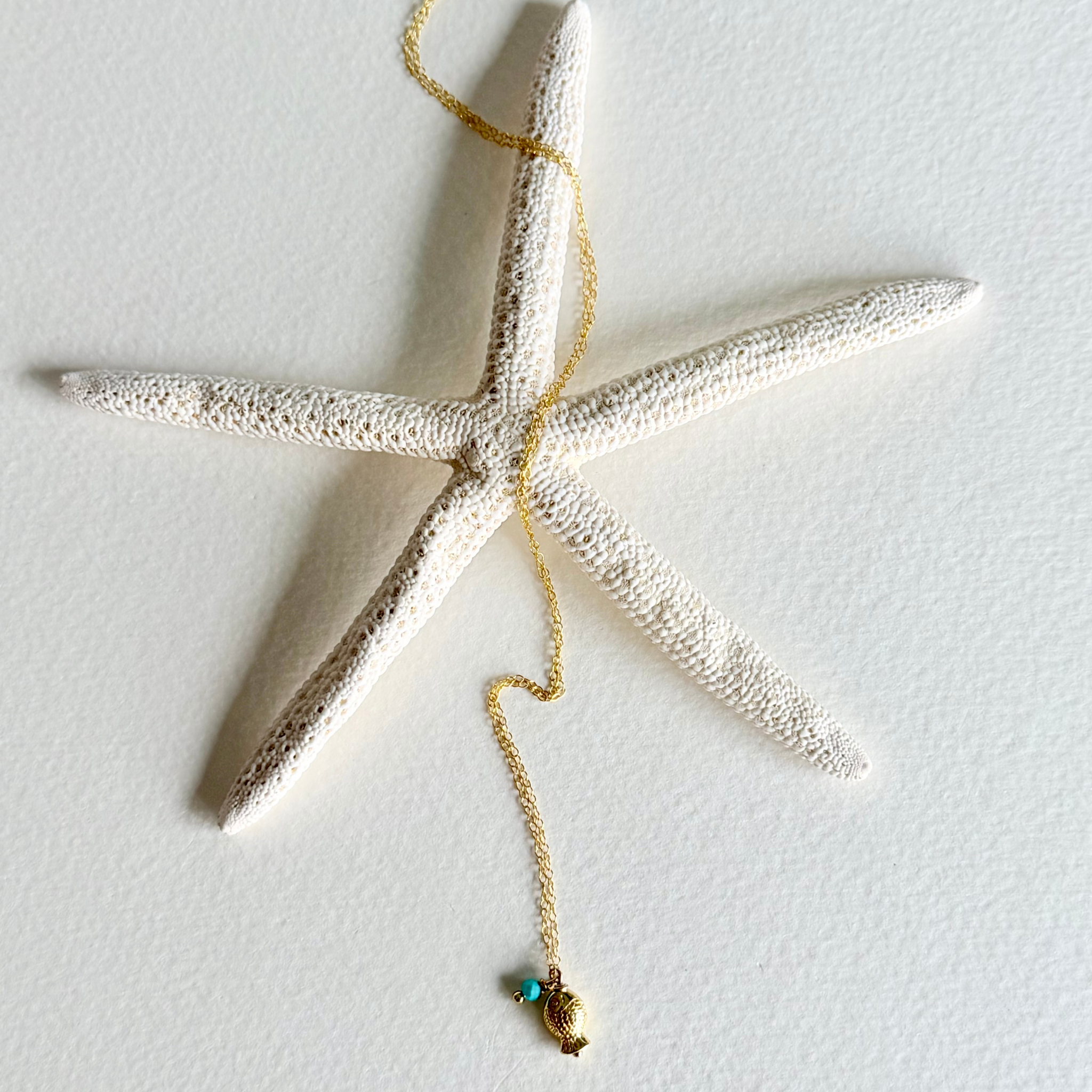 A textured white starfish wall hanging with a gold-toned chain draped over it and a small shell and turquoise pendant at the end, shown against a plain white wall.